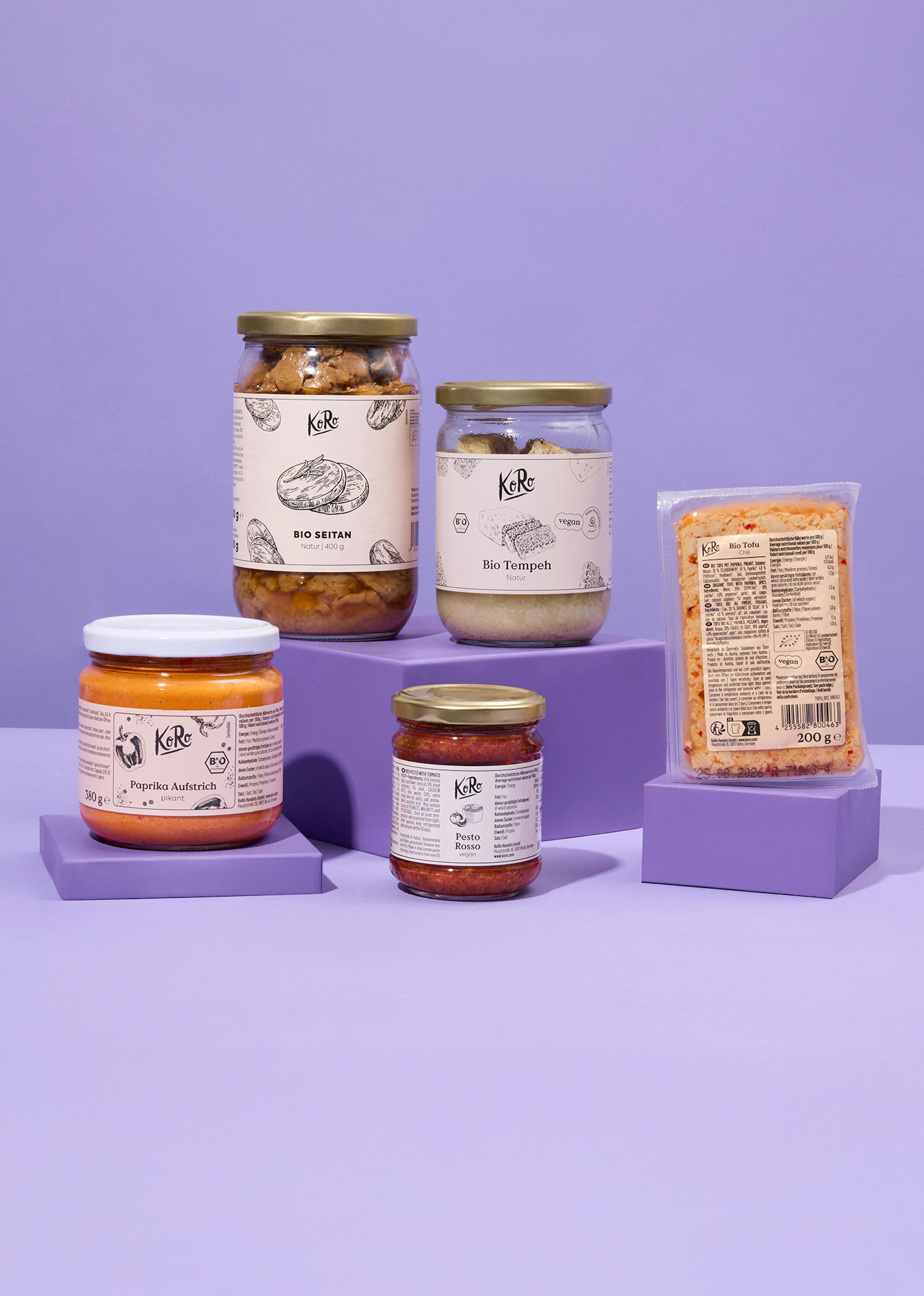 cinque prodotti alimentari vegani koro, tra cui seitan, tempeh, tofu, crema di paprika e pesto rosso, sono esposti su supporti viola su uno sfondo viola.