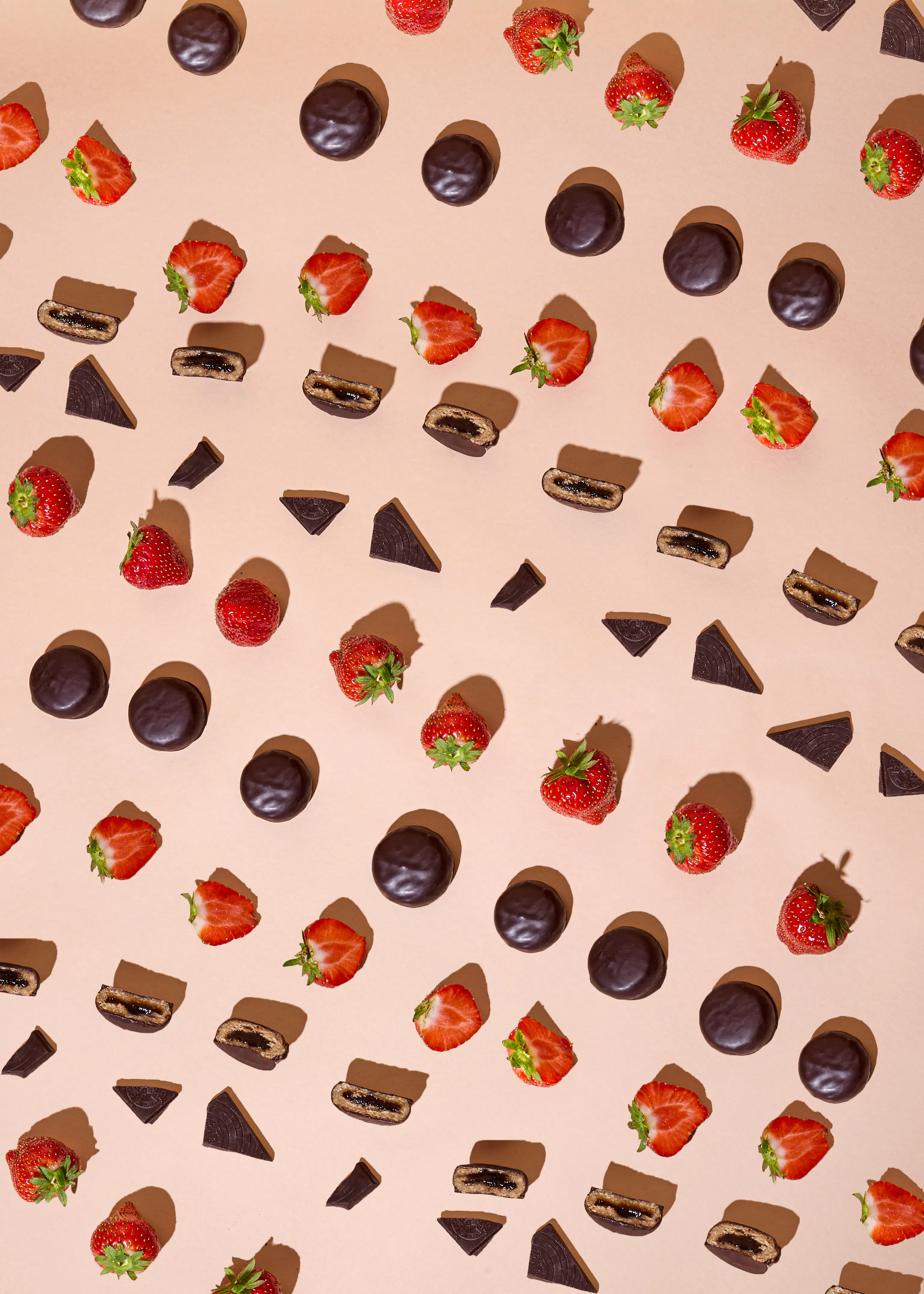 ein muster aus ganzen und halbierten erdbeeren, schokoladenkeksen und schokoladendreiecken auf einem hellrosa hintergrund mit schatten.