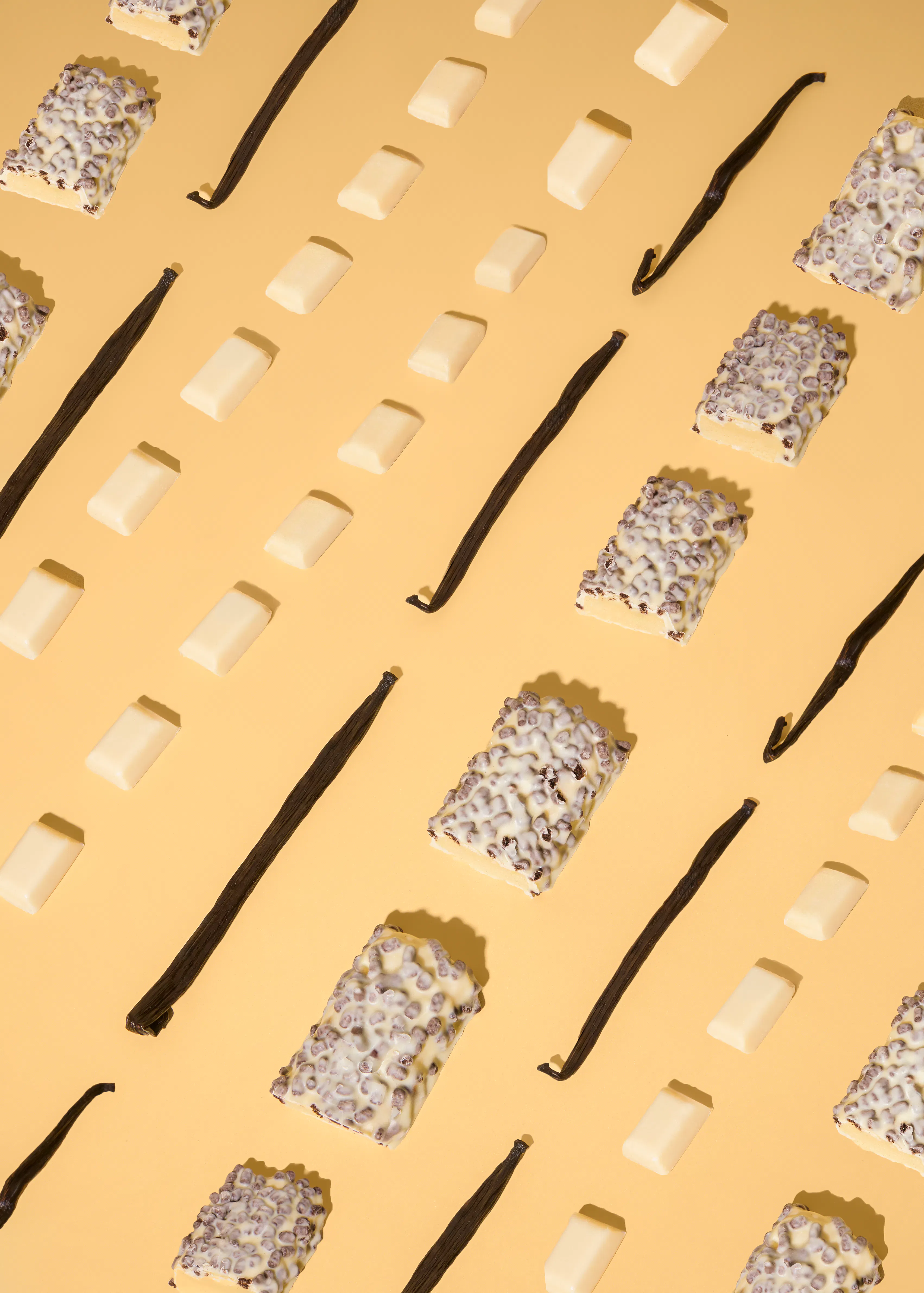 un motivo diagonale di barrette di cioccolato bianco, baccelli di vaniglia e barrette di cioccolato bianco con granella scura su uno sfondo giallo chiaro.
