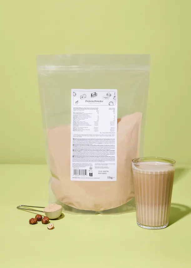 sac de poudre protéinée kore vegan saveur noisette crémeuse de 1 kg, un verre de boisson protéinée, une cuillère doseuse avec de la poudre et des noisettes sur un fond vert clair.