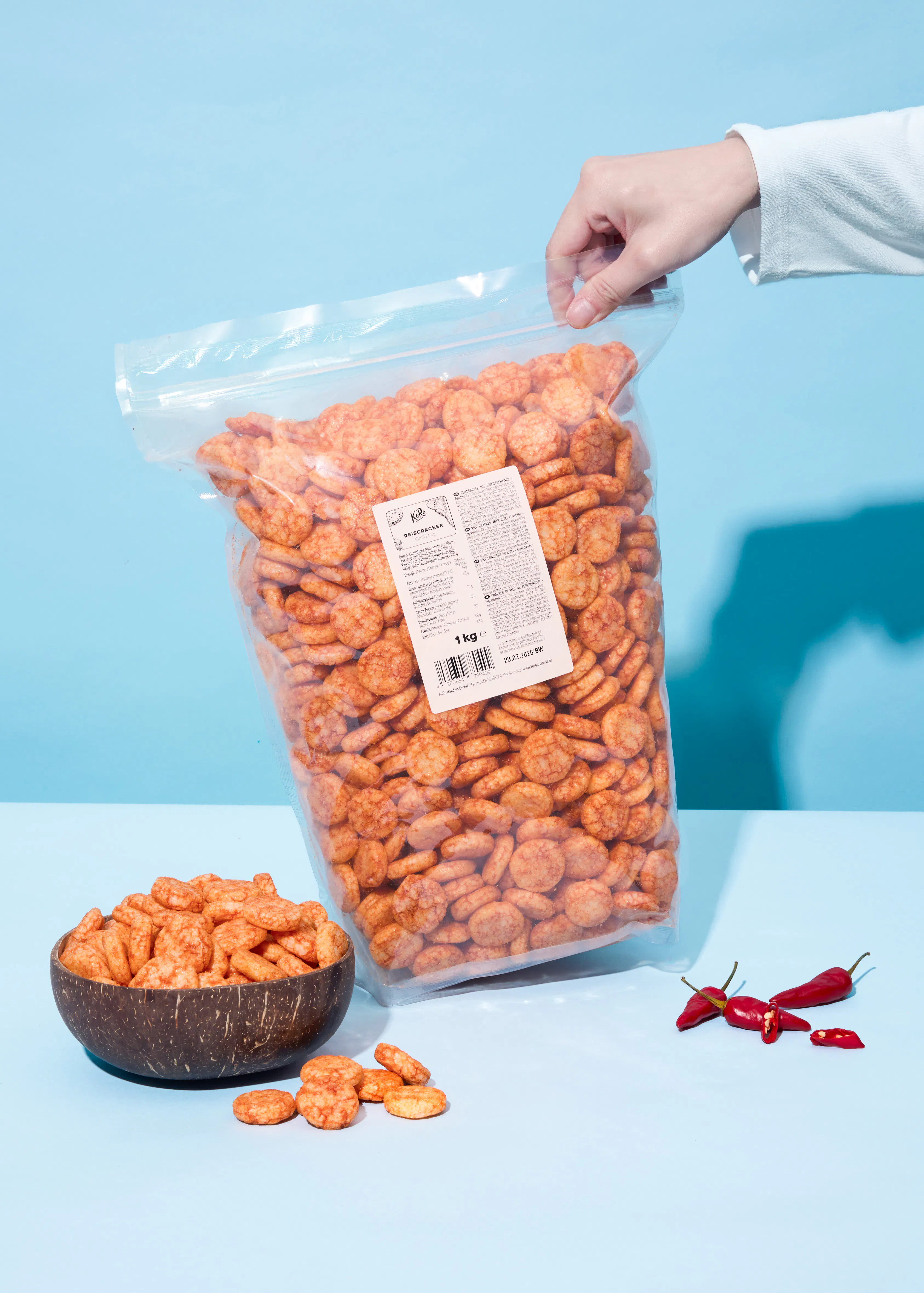 une main tient un grand sac transparent de crackers de riz orange, avec un bol de crackers et des piments rouges sur une surface bleue.