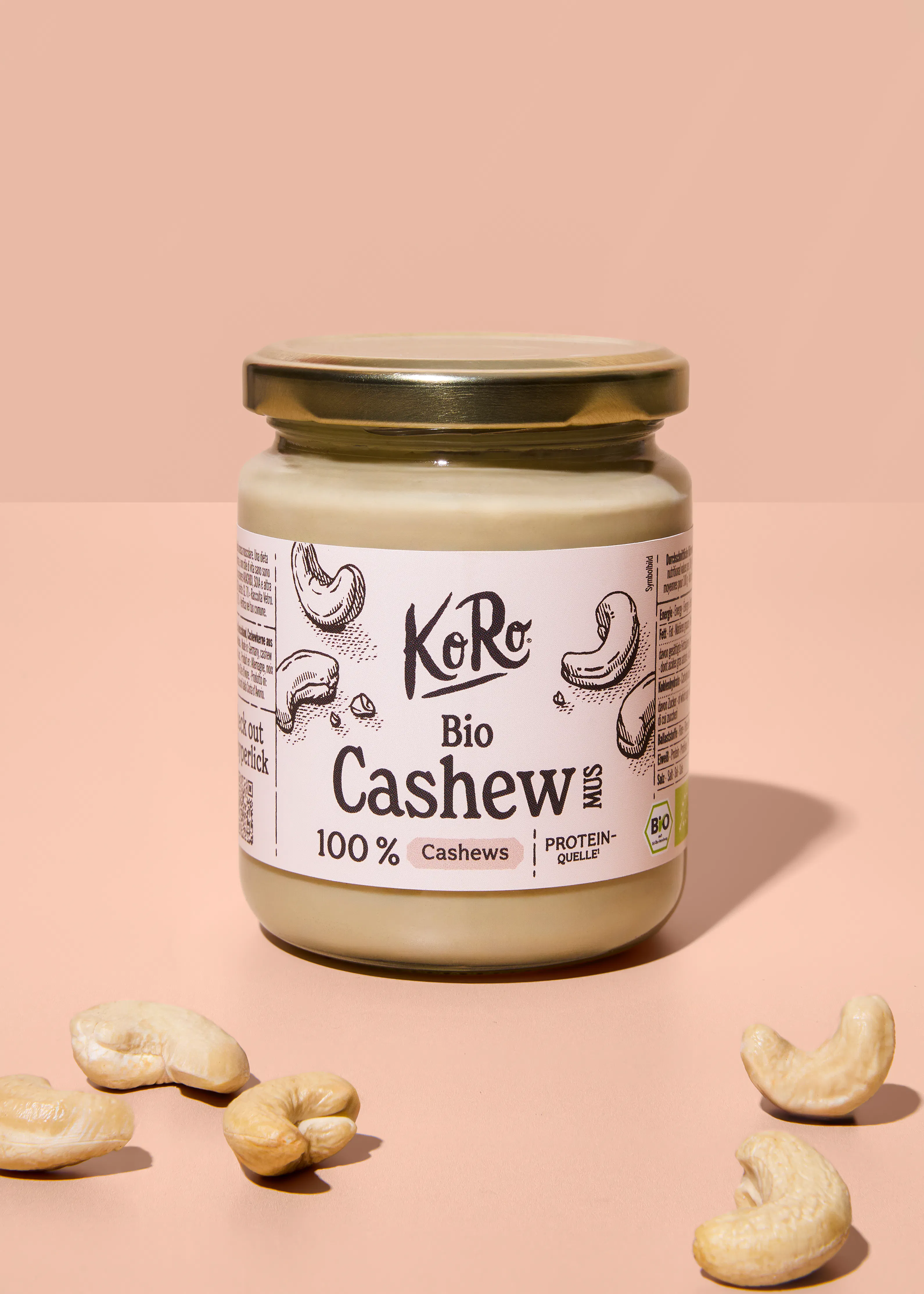 ein glas koro bio cashewmus mit goldenem deckel, umgeben von mehreren cashewkernen, auf einem hellrosafarbenen hintergrund.