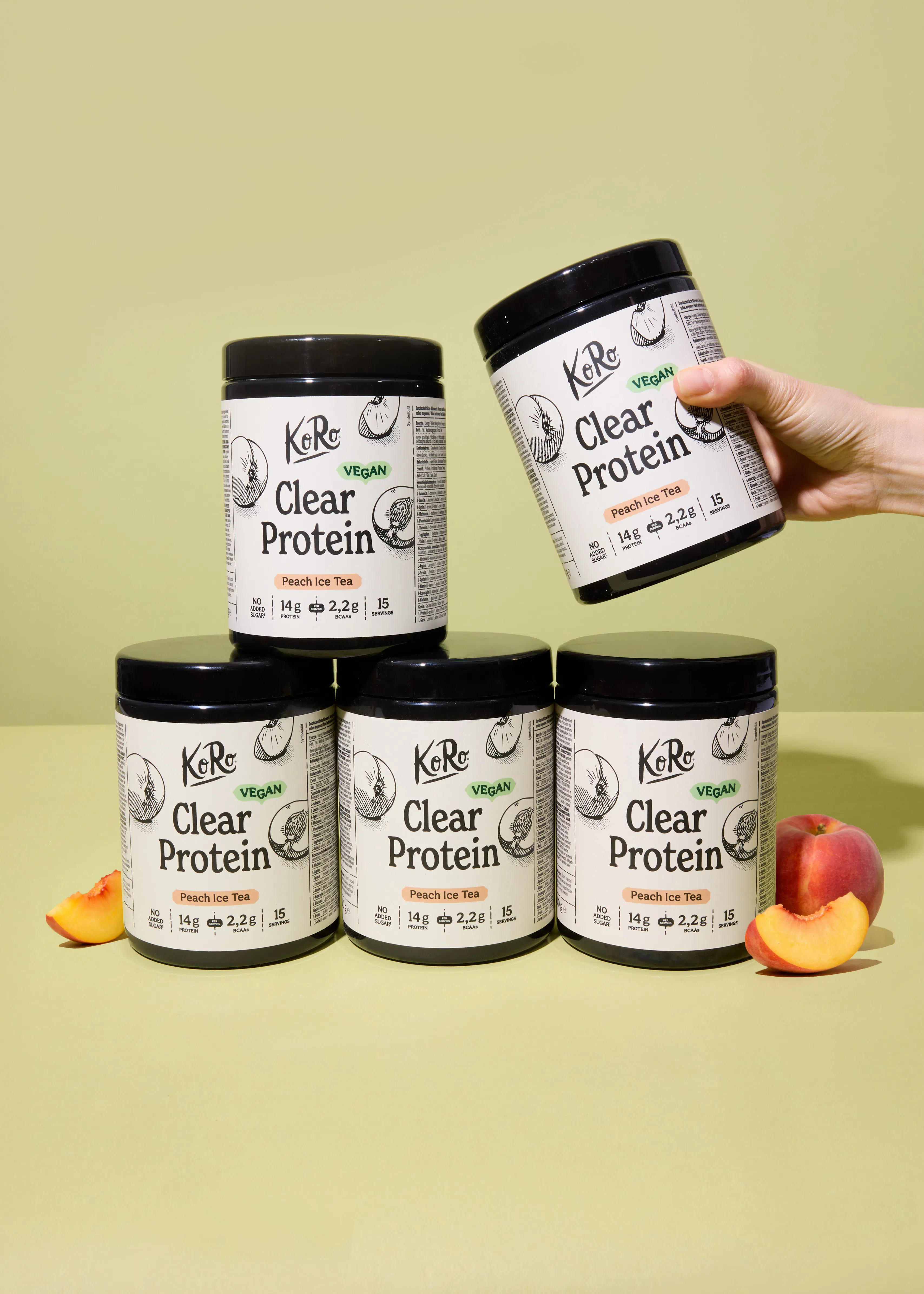 a hand holds one black koro clear protein peach ice tea container above four others, with fresh peaches and slices on a light green background.