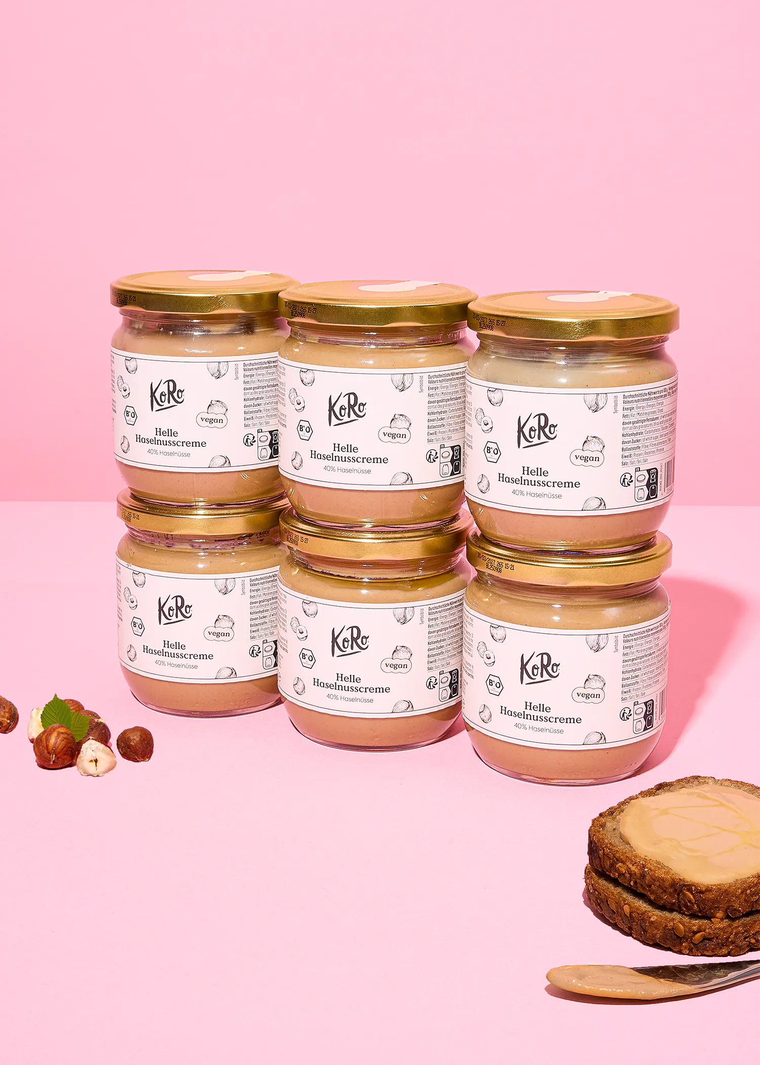 six pots de crème de noisette koro, des noisettes et une tranche de pain tartinée sont disposés sur un fond rose.