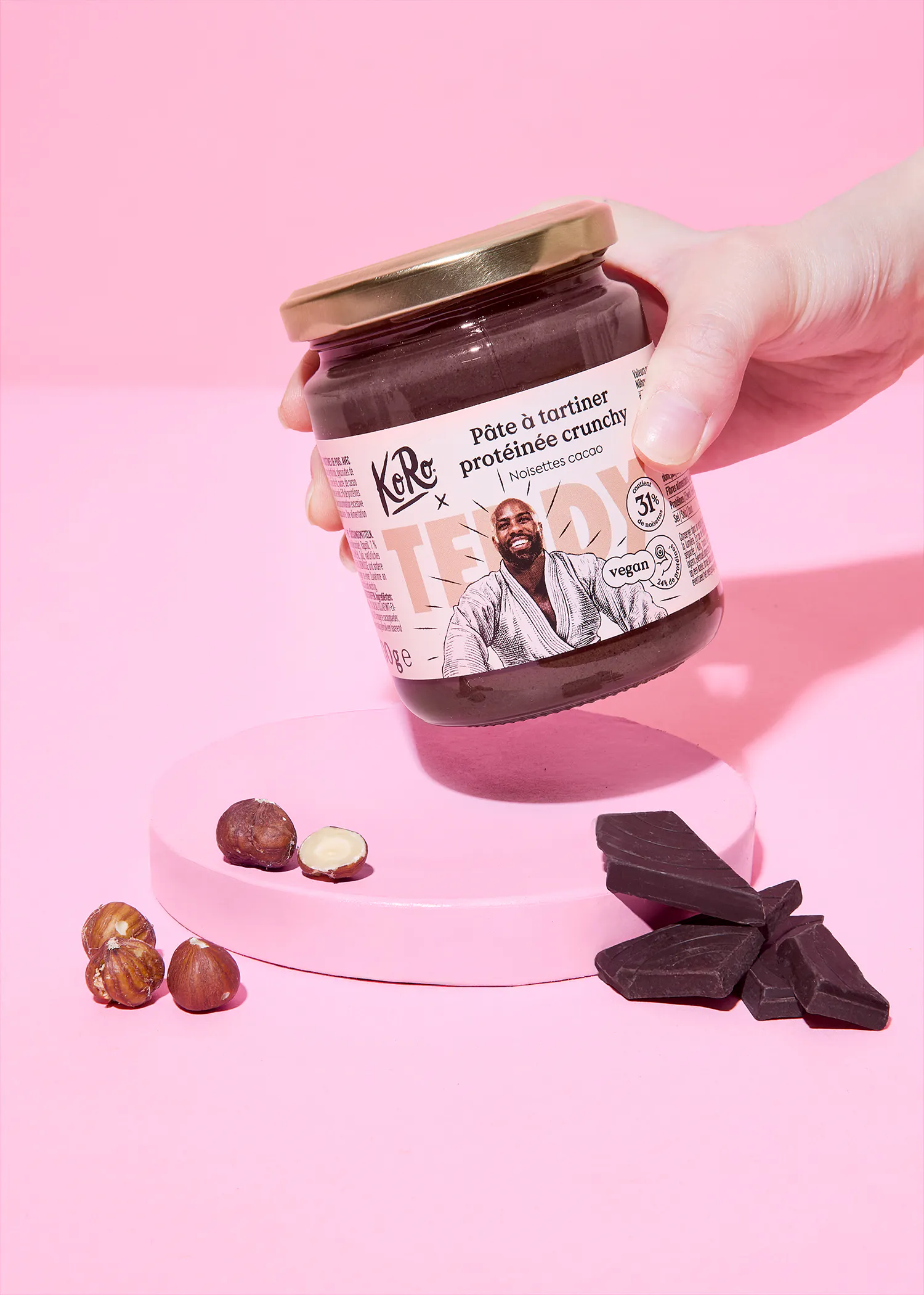 une main tient un pot de pâte à tartiner protéinée crunchy noisettes cacao koro avec l'illustration de teddy riner, au-dessus d'une plateforme rose avec des noisettes et des morceaux de chocolat noir.