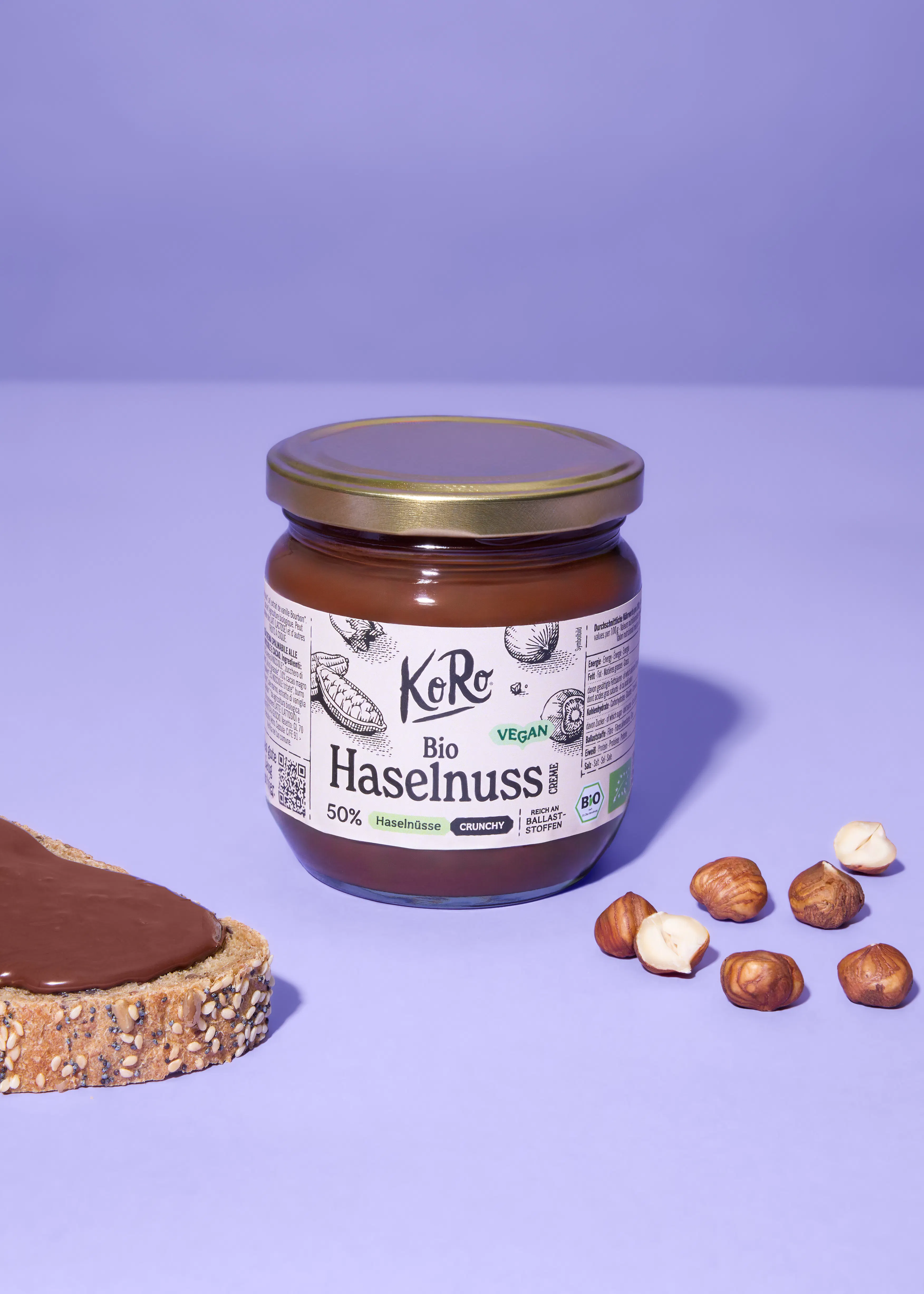 ein glas koro bio haselnusscreme mit goldfarbenem deckel steht neben einem mit haselnusscreme bestrichenen brot und verstreuten ganzen und halbierten haselnüssen auf einem lila hintergrund.