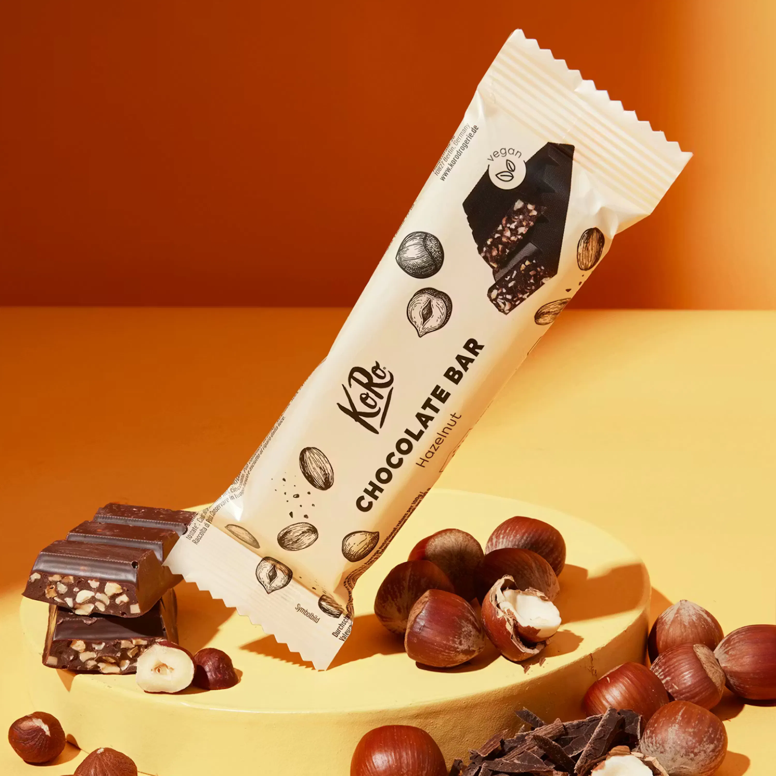 una barretta di cioccolato koro alle nocciole, parzialmente aperta per mostrare il cioccolato con pezzi di nocciola, è appoggiata su una superficie gialla con nocciole intere e pezzi di cioccolato sparsi intorno, su uno sfondo arancione.