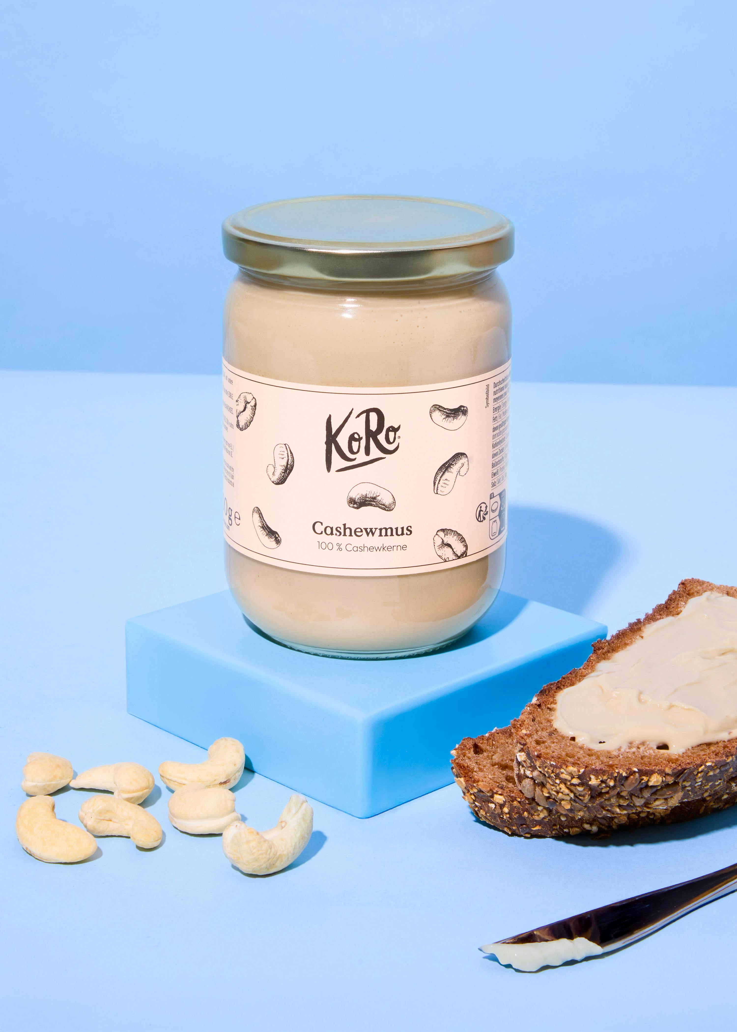 ein glas cashewmus, lose cashews und eine scheibe brot mit cashewmus auf blauem hintergrund.