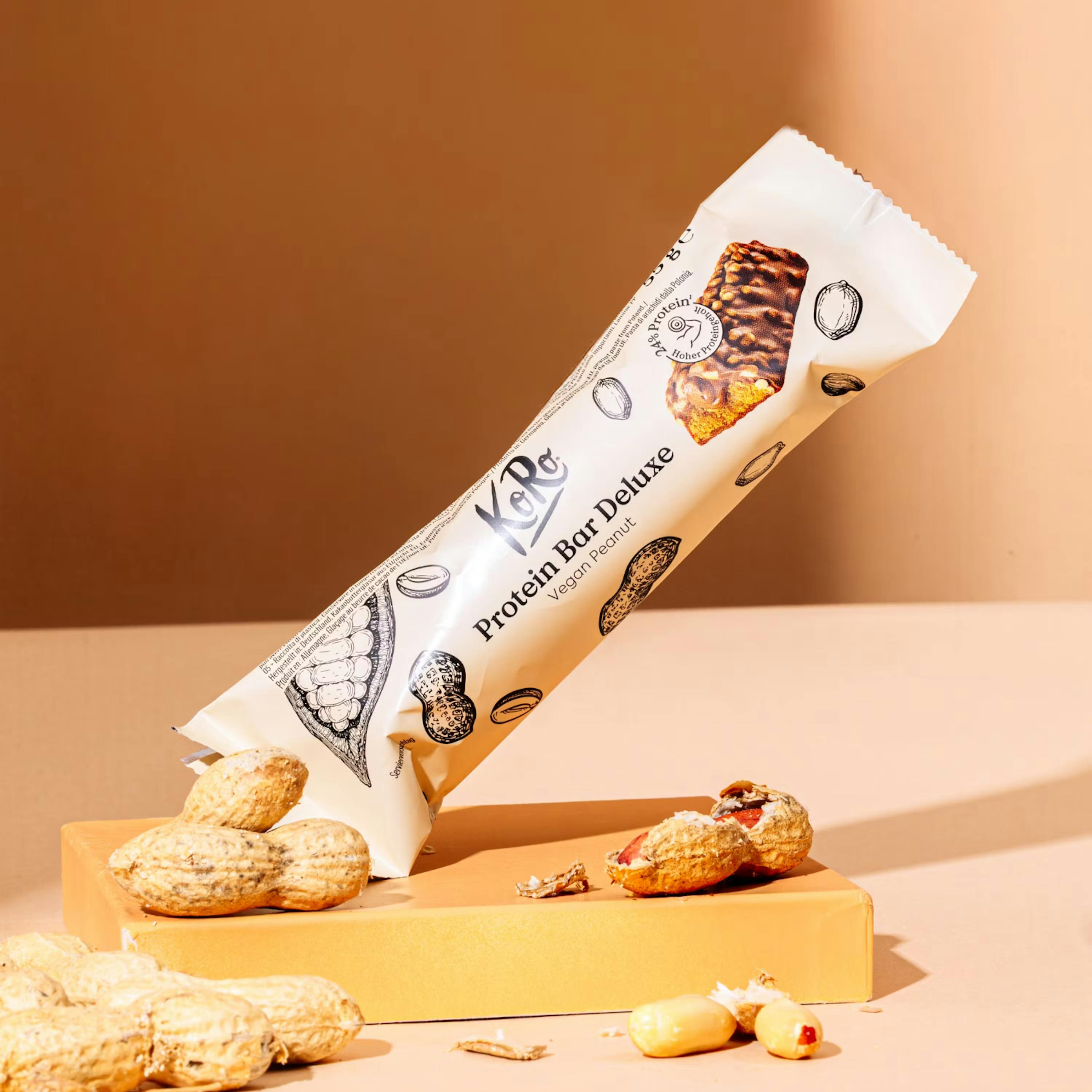 a koro protein bar deluxe vegan peanut is partially unwrapped on an orange surface with peanuts scattered around it.