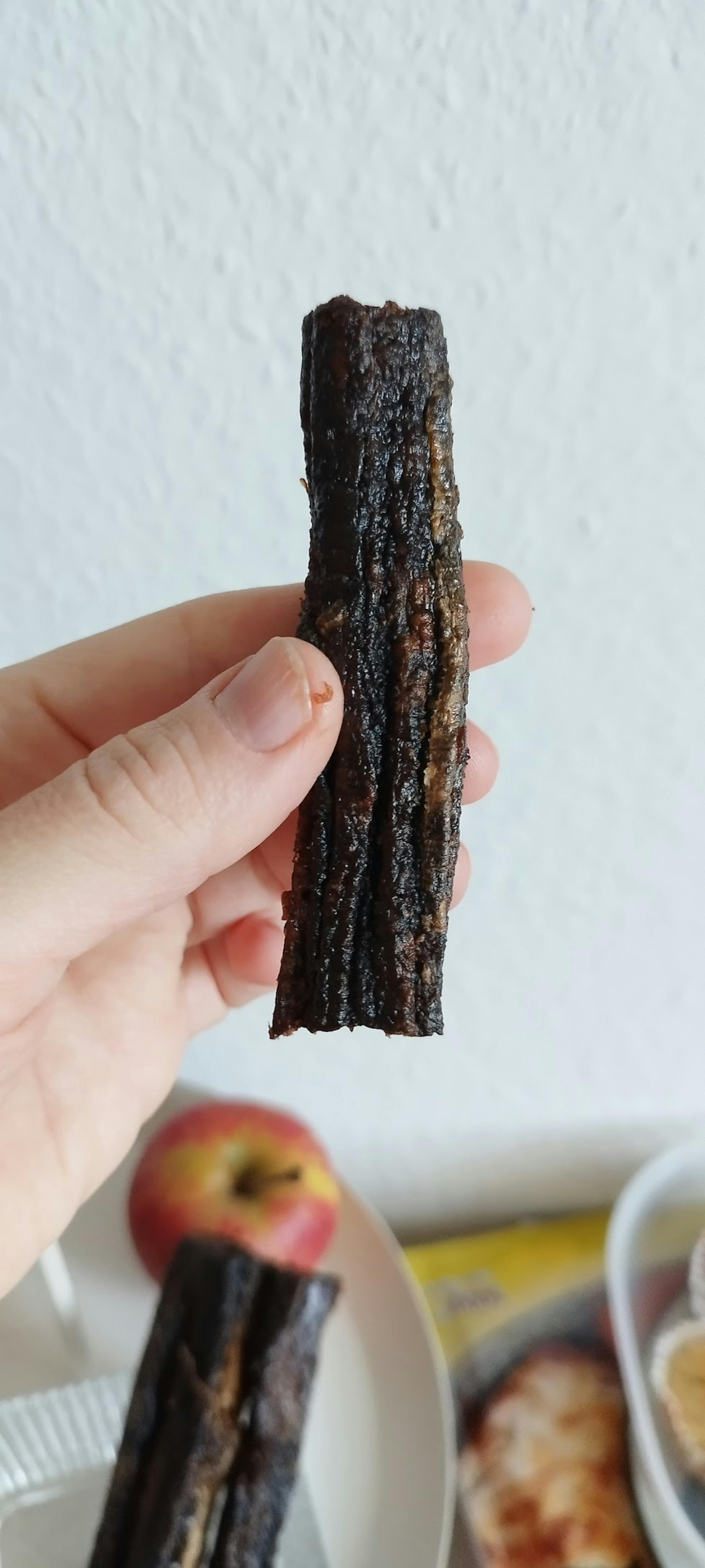 a hand holds a dark, textured, rectangular food stick against a light background.