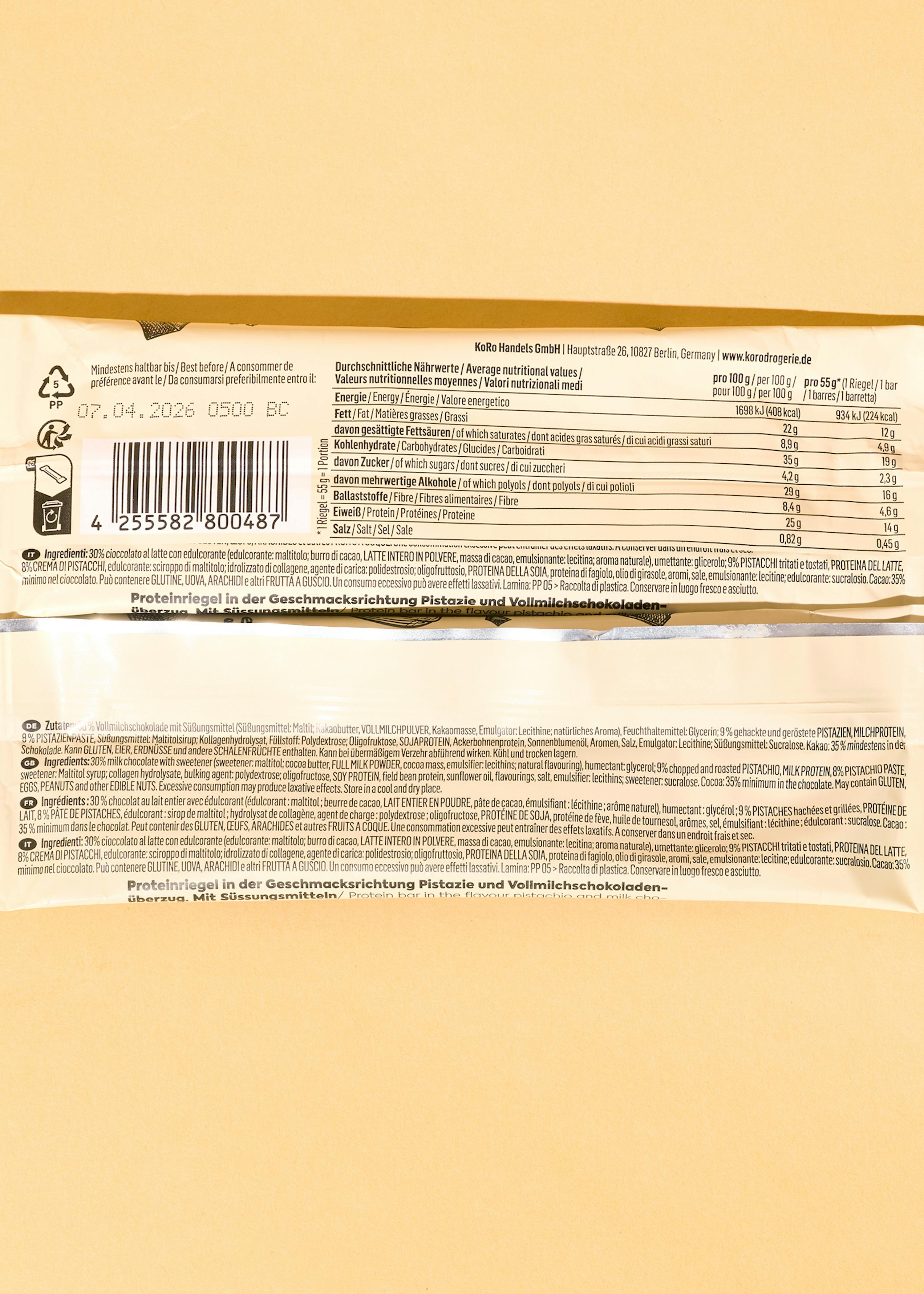 die rückseite einer verpackung für einen proteinriegel mit pistazien- und vollmilchschokoladengeschmack, die nährwertangaben, zutatenlisten in mehreren sprachen, ein haltbarkeitsdatum vom 07.04.2026 und einen strichcode zeigt.