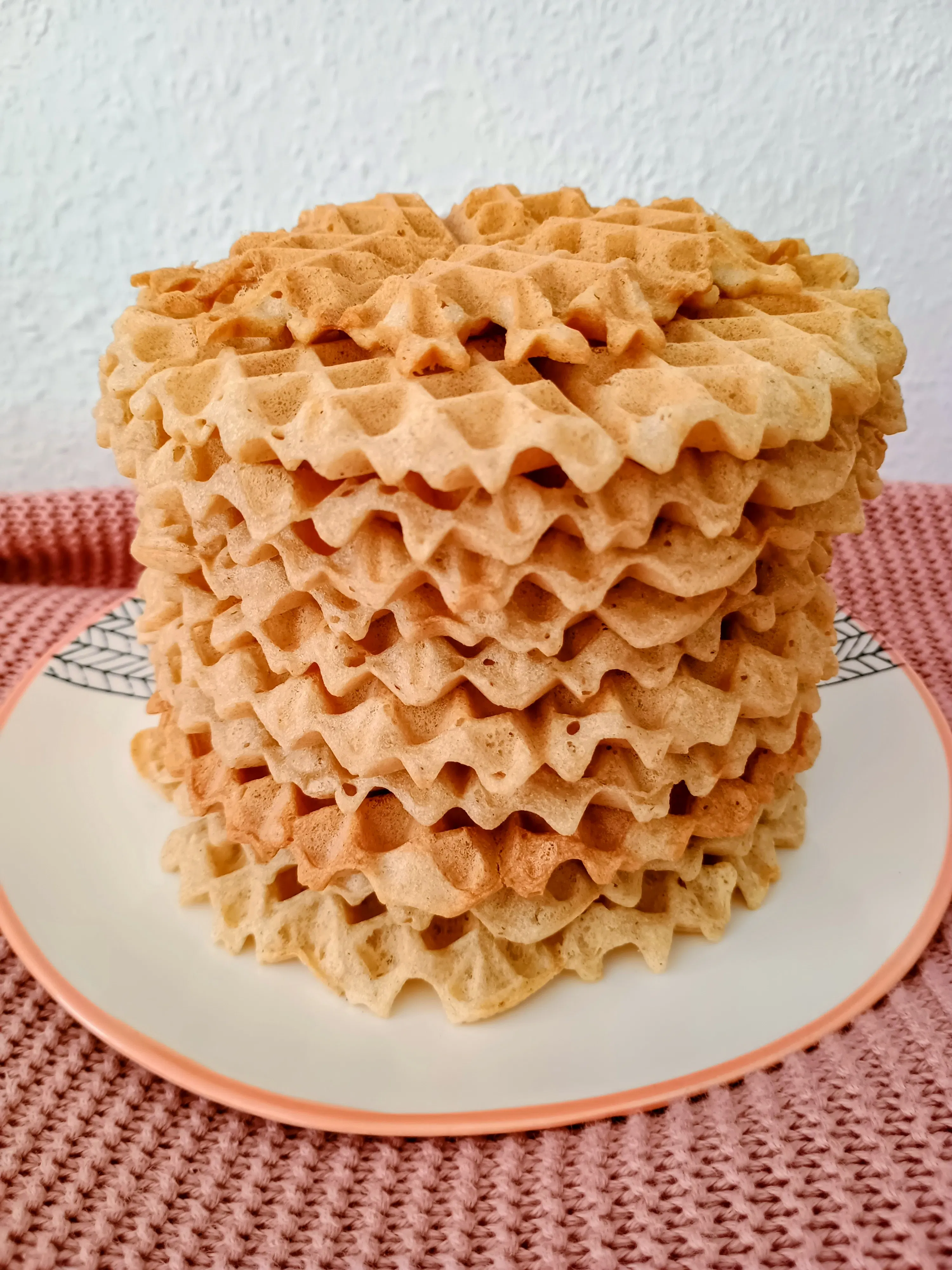 a tall stack of golden-brown, wavy waffles on a white plate with a pink rim, sitting on a pink knitted surface.