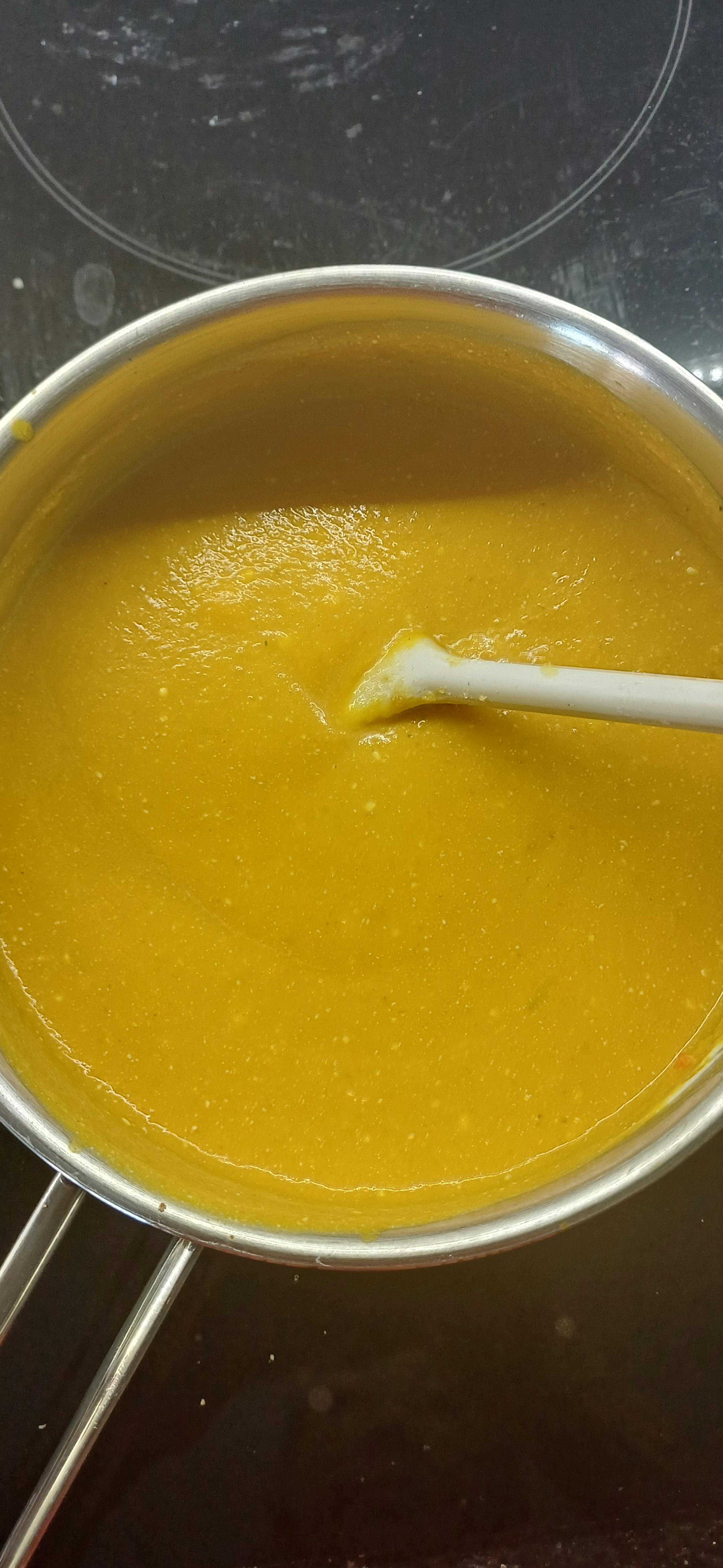a top-down view of a metal pot filled with thick, yellow-orange soup or sauce, with a white spoon resting in it, on a dark stovetop.