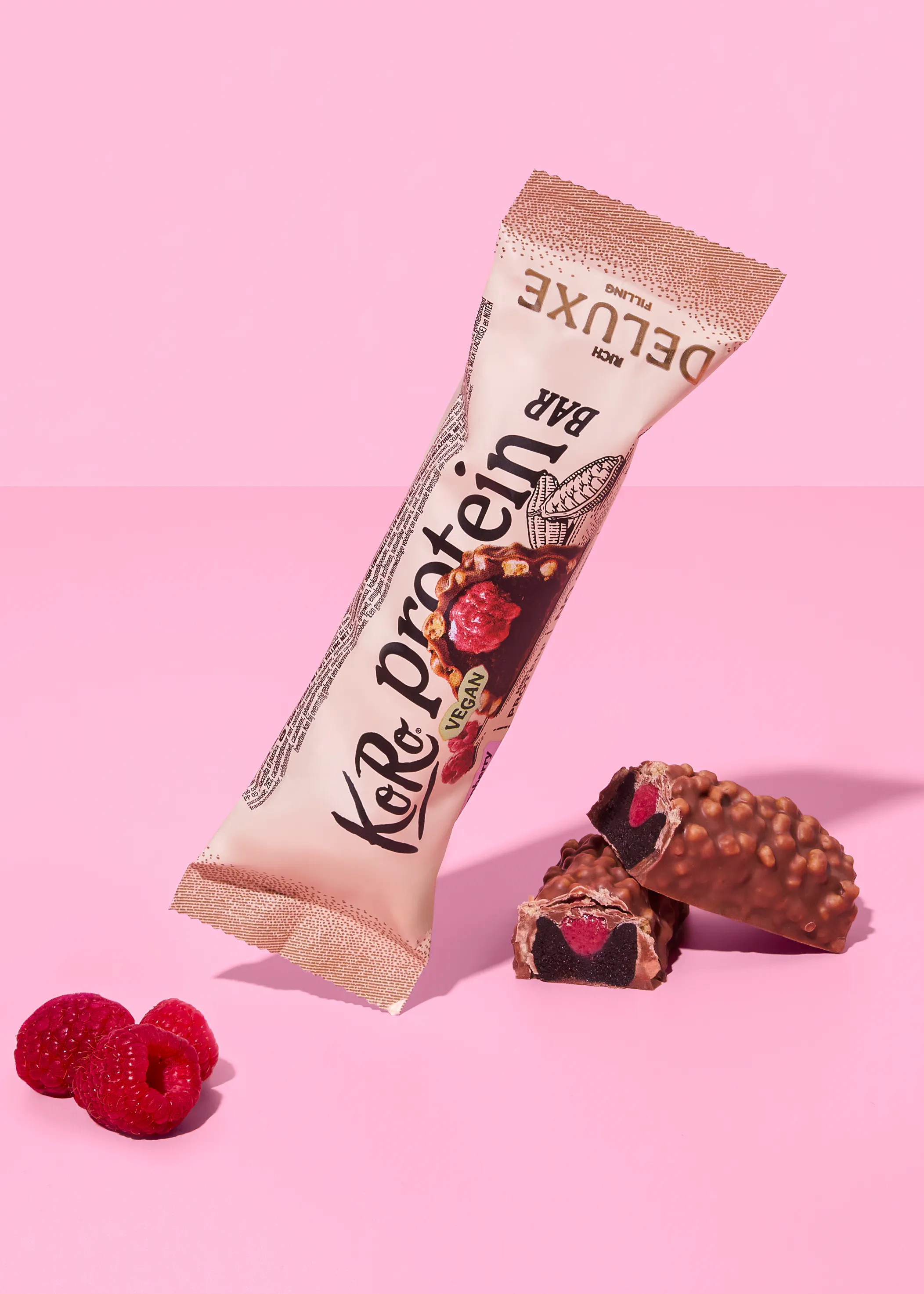 eine koro protein deluxe riegelverpackung, ein zerbrochener riegel mit dunkler füllung und rotem kern sowie frische himbeeren auf rosa hintergrund.