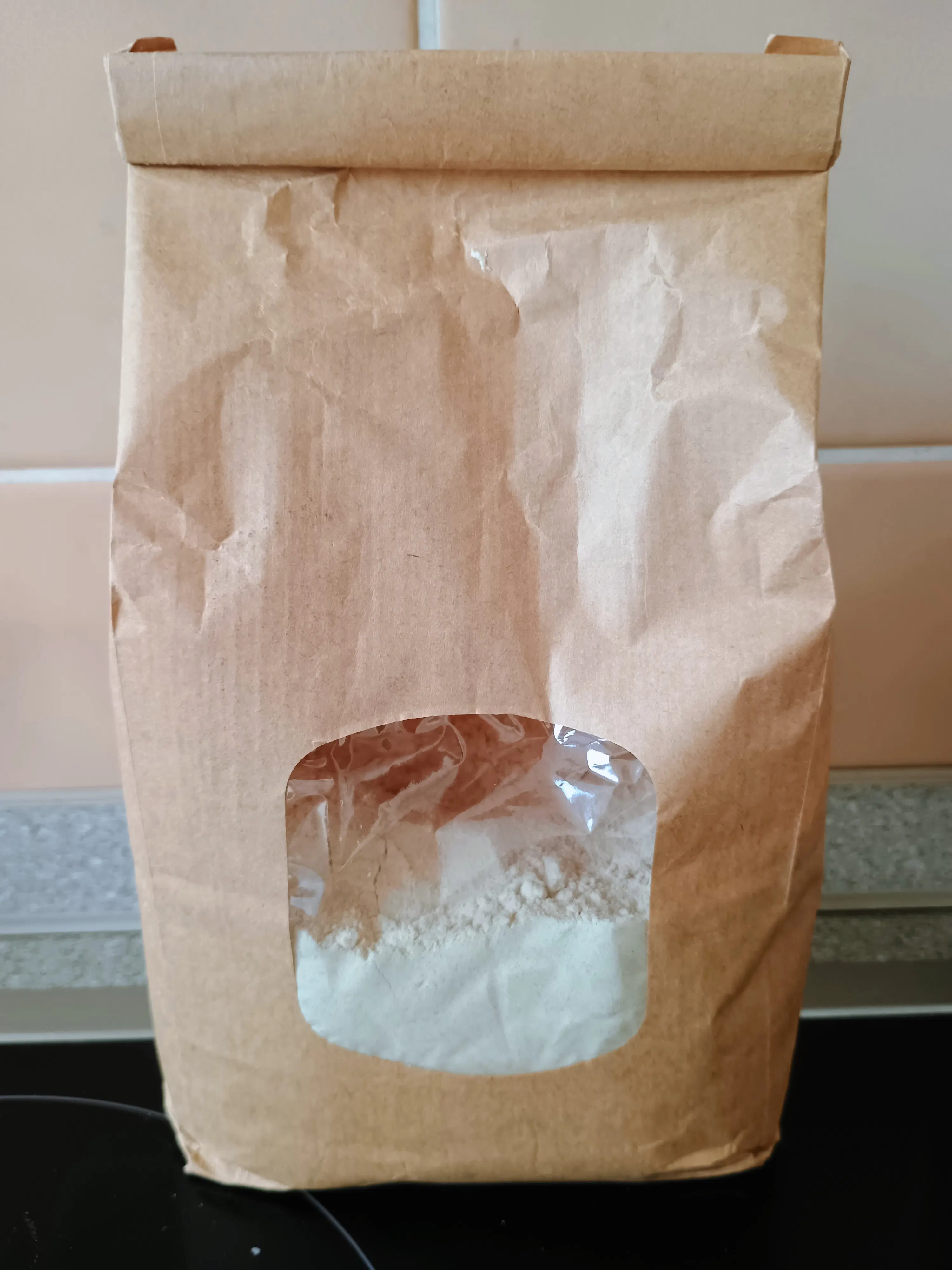 a brown paper bag with a clear window showing white flour inside stands on a counter.