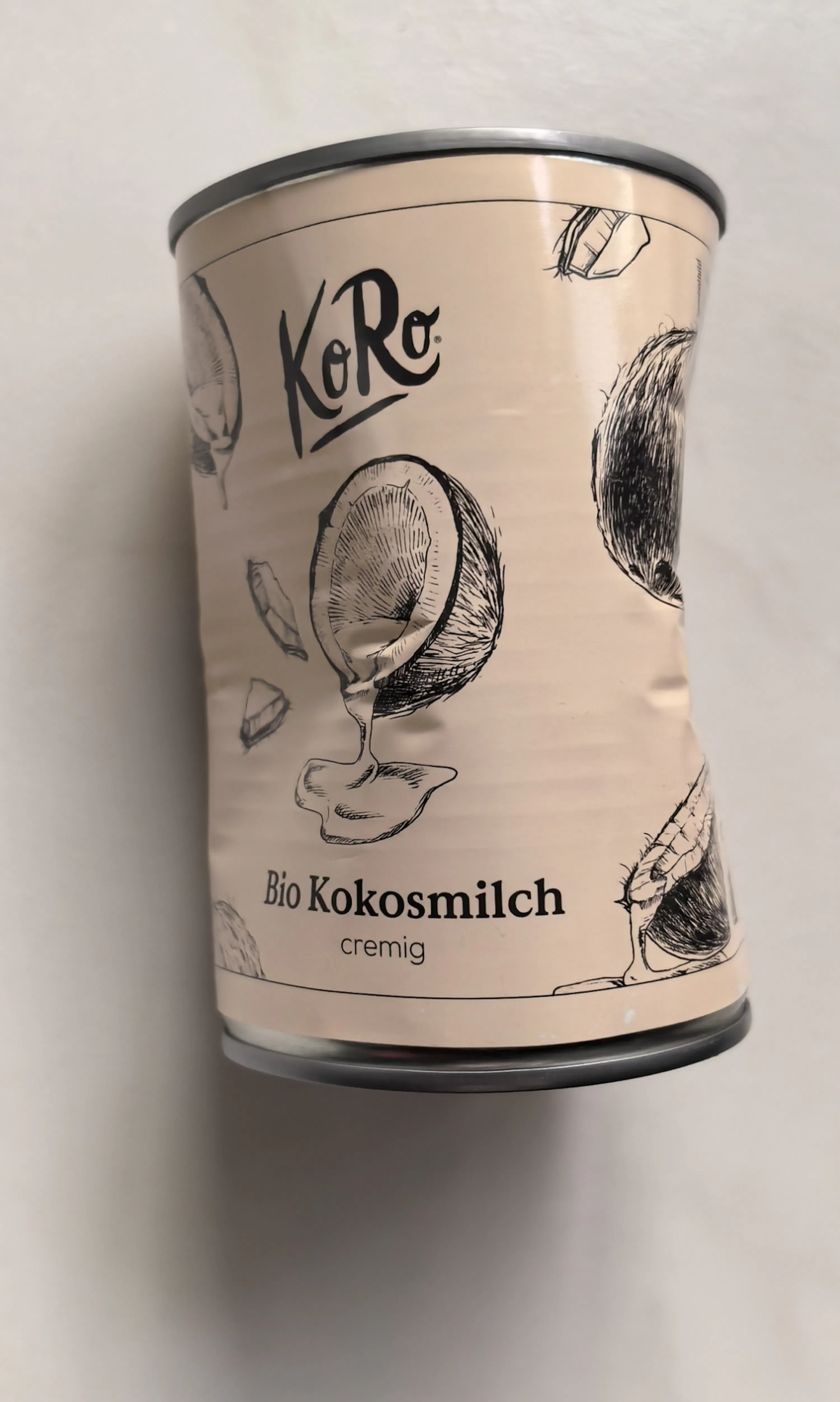 une boîte de lait de coco bio koro crémeux, légèrement bosselée, avec des illustrations de noix de coco et de lait de coco versé sur une étiquette beige clair.