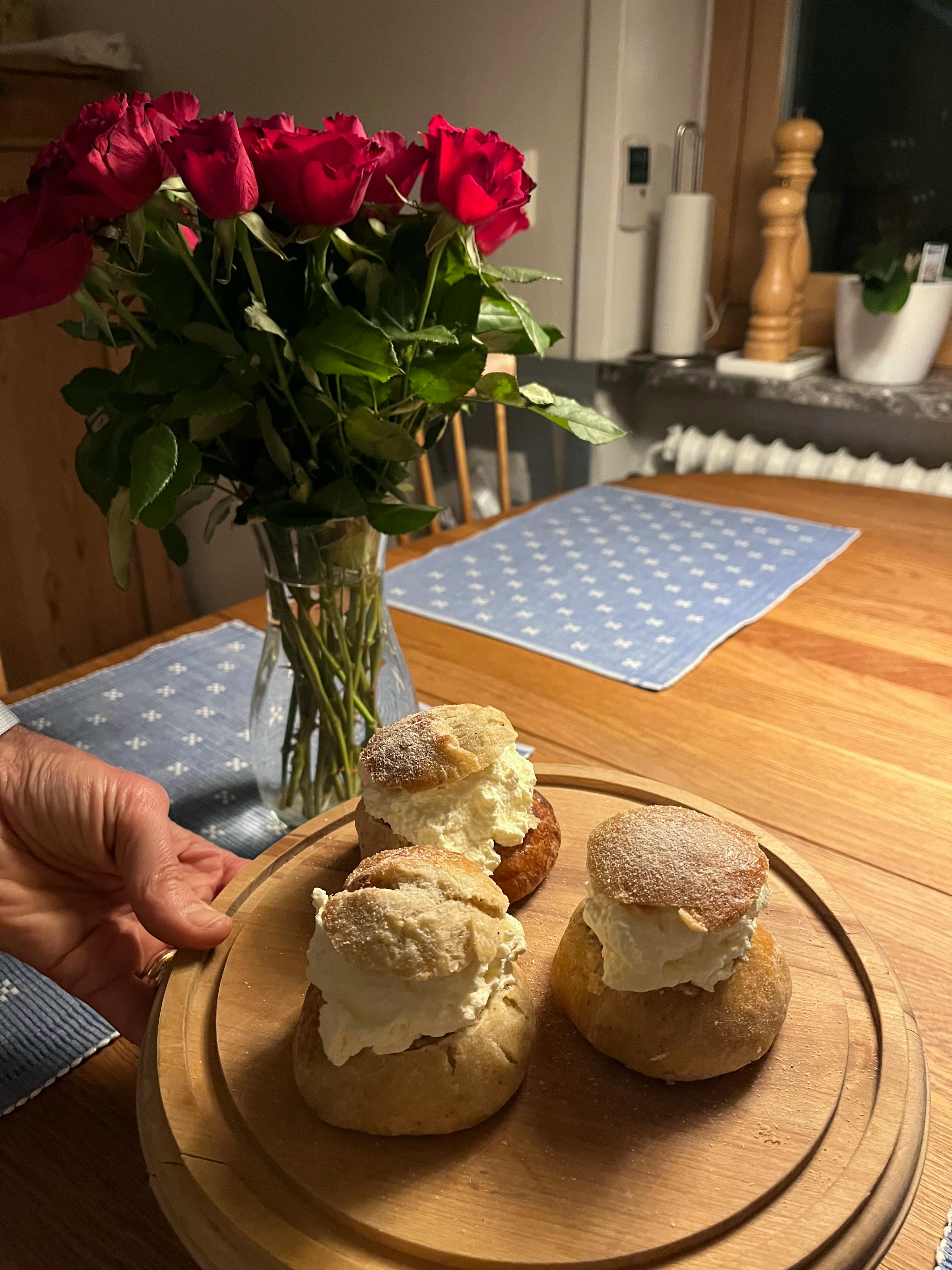 a hand holds a wooden serving board with three cream-filled buns, next to a vase of red roses on a wooden table.