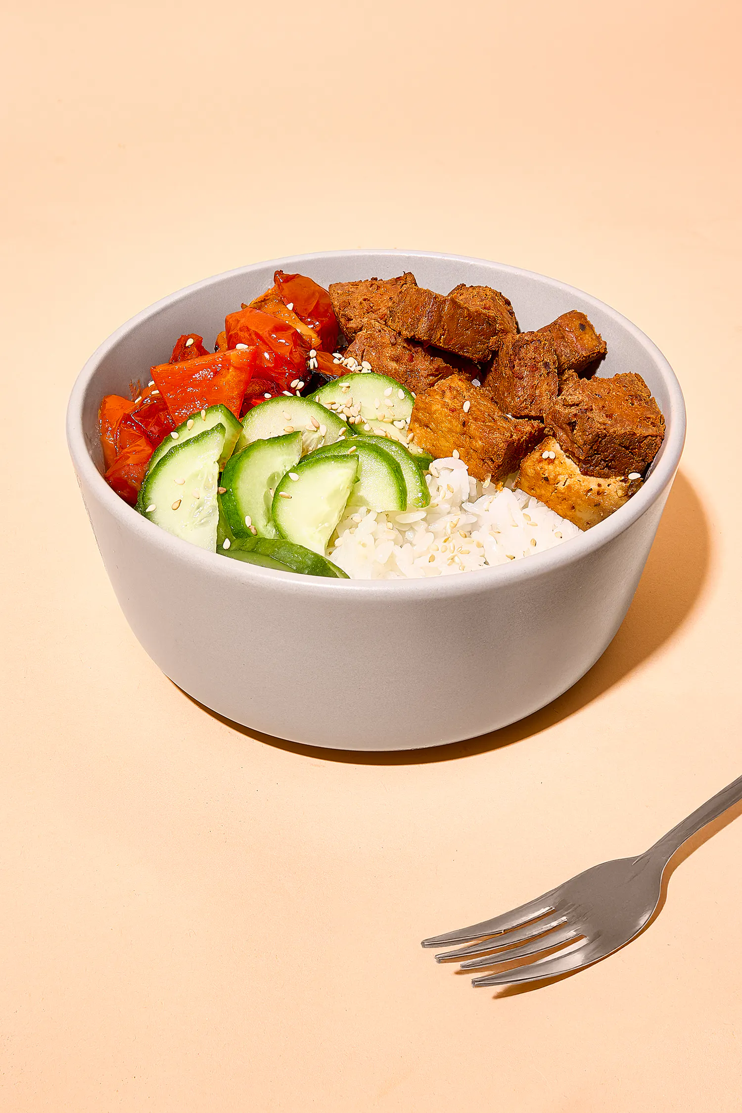 un bol de riz blanc garni de concombres tranchés, de poivrons rouges rôtis, de cubes de tofu assaisonnés et de graines de sésame, posé sur une surface beige avec une fourchette.