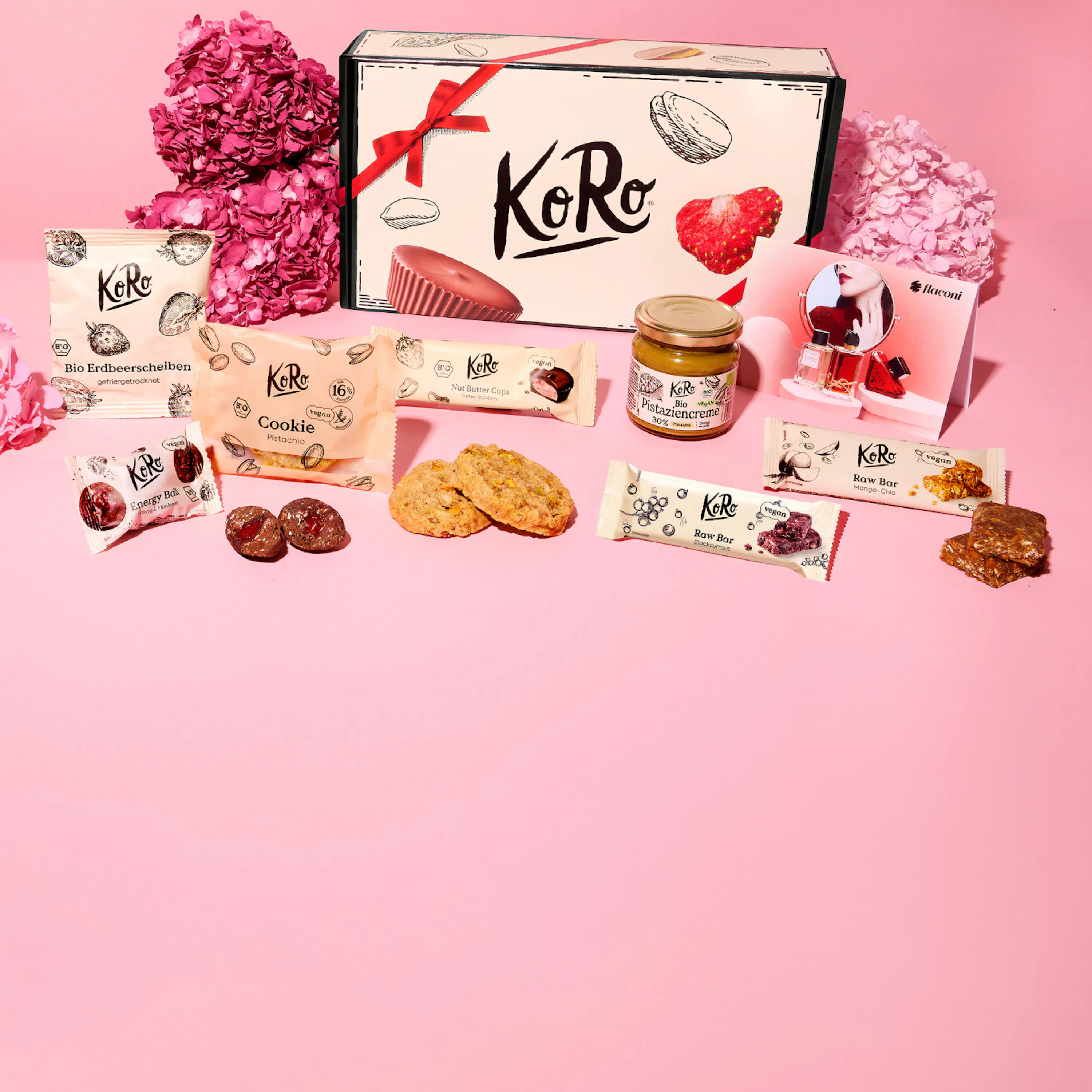 eine festliche anordnung von koro-snacks wie keksen, riegeln und cremes, zusammen mit einer geschenkbox und rosa blumen auf einem rosa hintergrund.