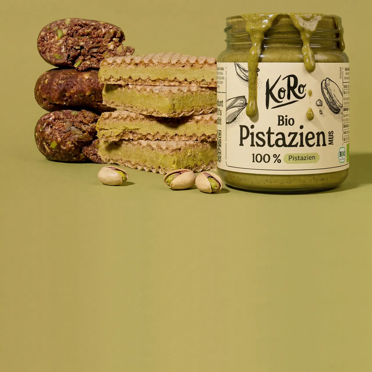 ein glas pistazienmus mit tropfen, daneben gestapelte pistazienwaffeln und energiekugeln sowie lose pistazien auf grünem hintergrund.