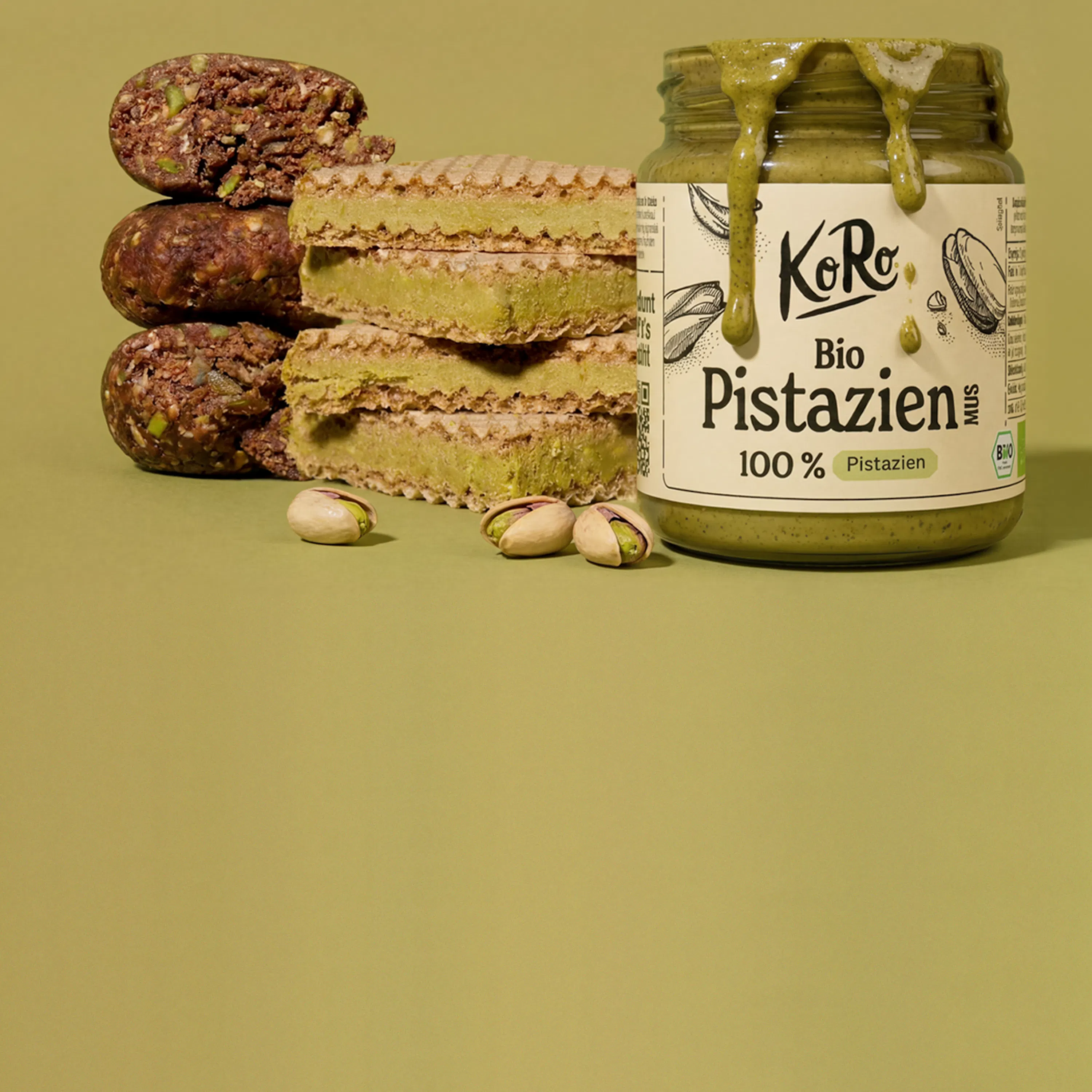 ein glas pistazienmus mit tropfen, daneben gestapelte pistazienwaffeln und energiekugeln sowie lose pistazien auf grünem hintergrund.