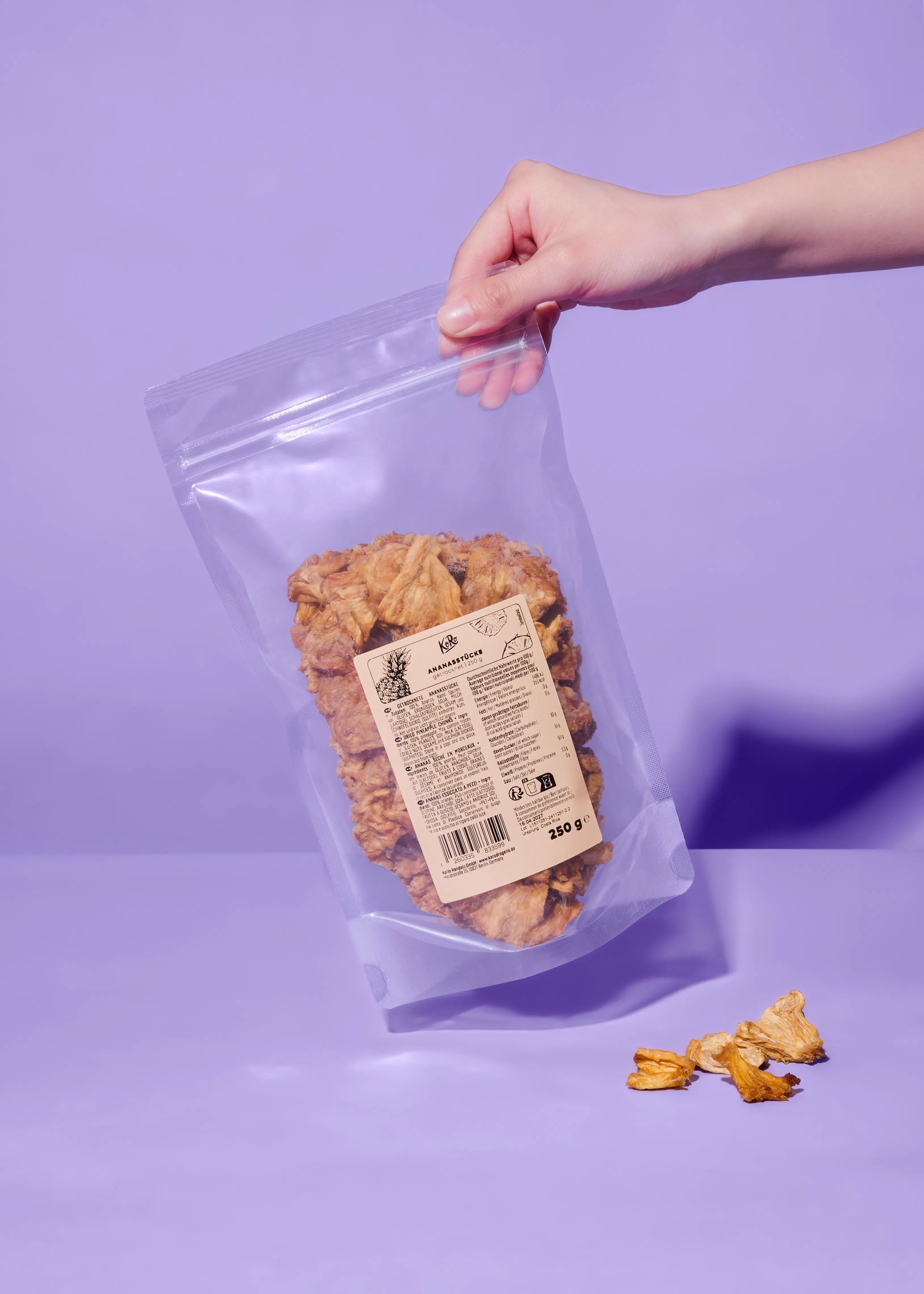 a hand holds a clear bag of dried pineapple pieces with a label showing "ananasstücke" and "250 ge", while a few pieces lie on the purple surface below.