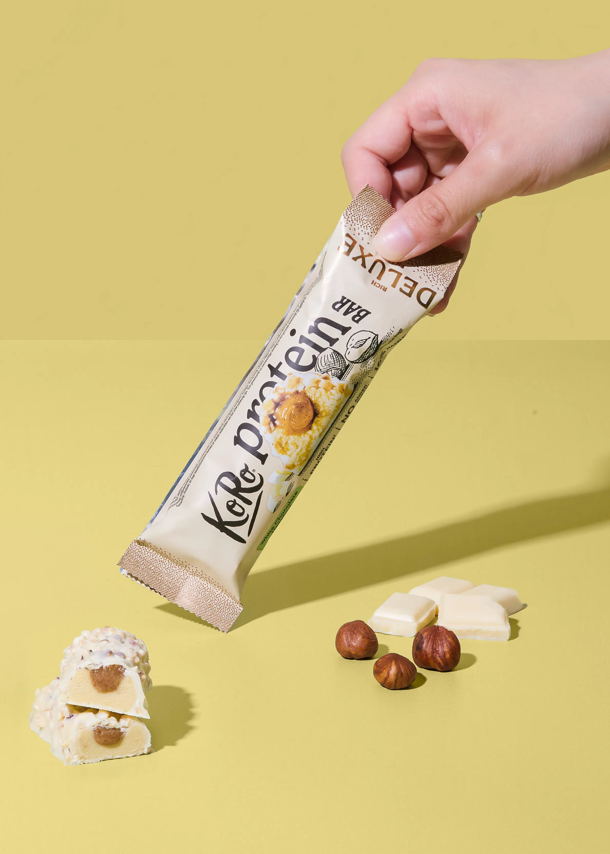 a hand holds a koro protein deluxe bar in white packaging with a hazelnut illustration, while two cut bar halves, three hazelnuts, and white chocolate pieces rest on a yellow surface.