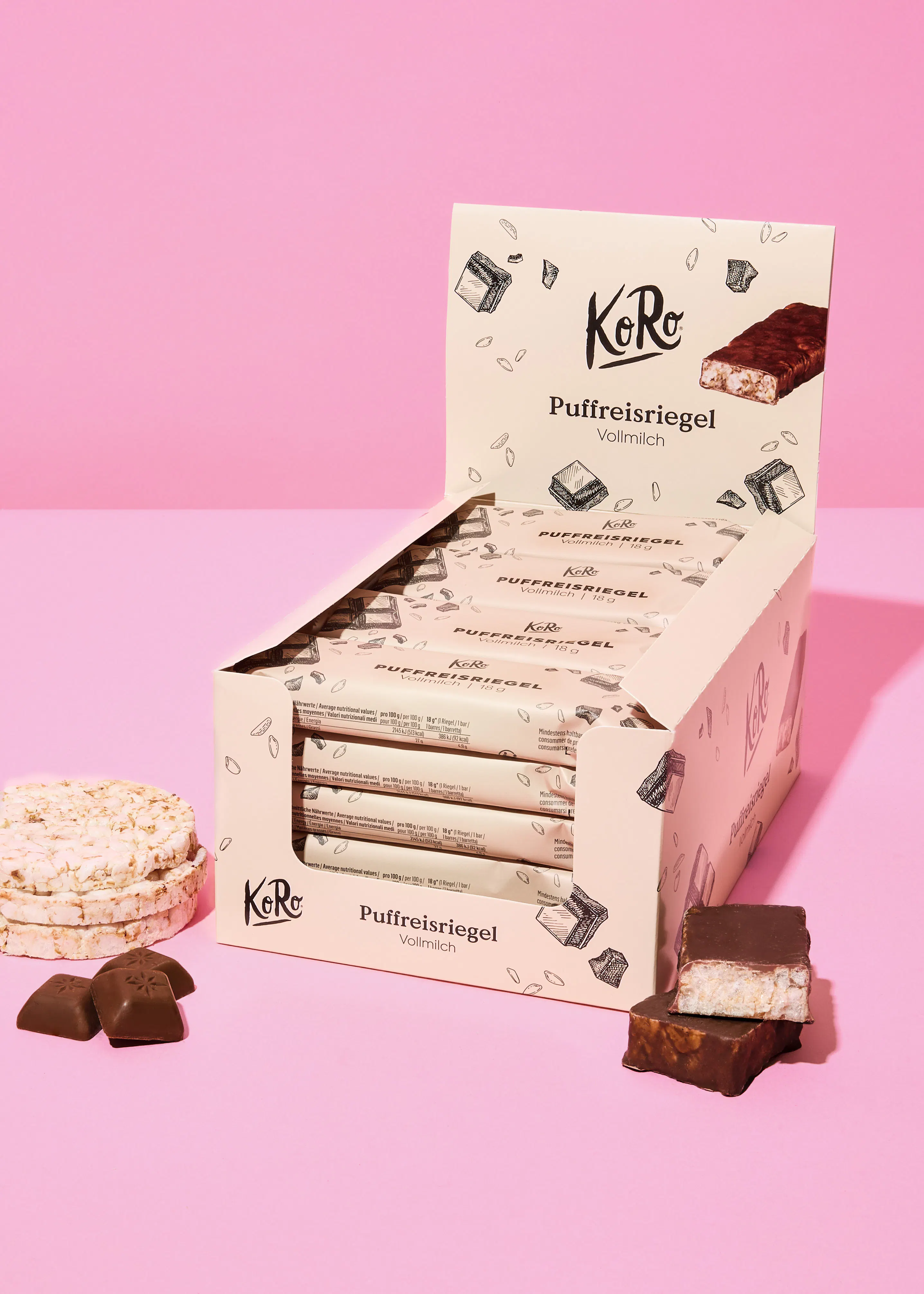eine offene schachtel mit mehreren koro puffreisriegeln vollmilch, ein gebrochener riegel, zwei schokoladenstücke und gestapelte reiswaffeln auf einem pinken hintergrund.