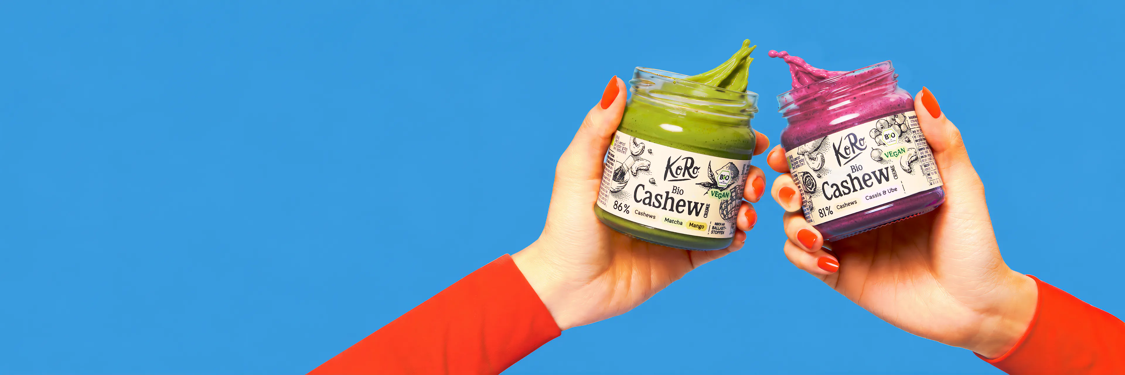 zwei hände halten vor blauem hintergrund je ein glas koro bio cashewcreme, eine grüne matcha-mango-creme und eine lila cassis-ube-creme, aus denen die creme herausquillt.