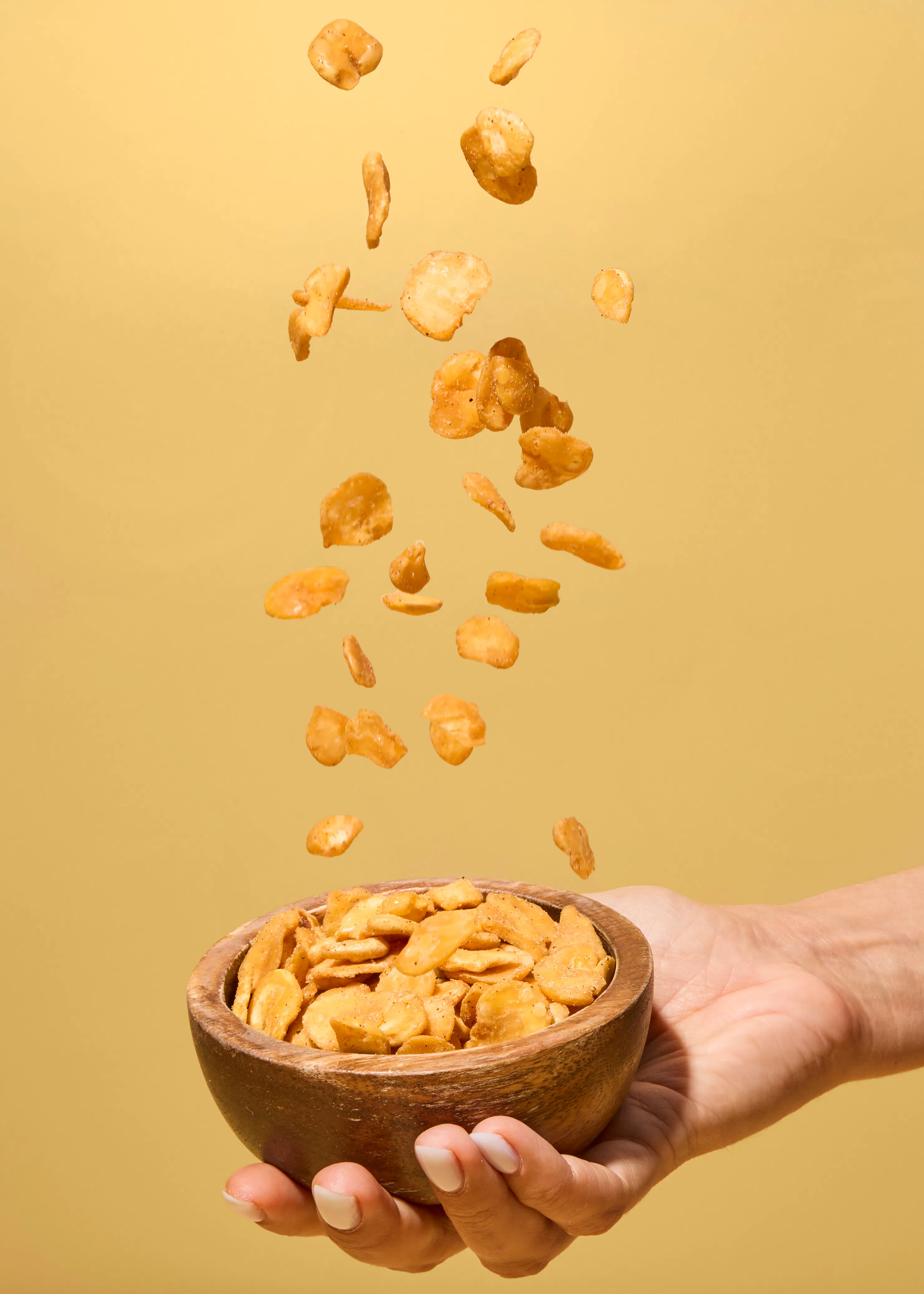 une main tient un bol en bois rempli de fèves frites alors que d'autres fèves tombent en suspension au-dessus du récipient sur un fond jaune.
