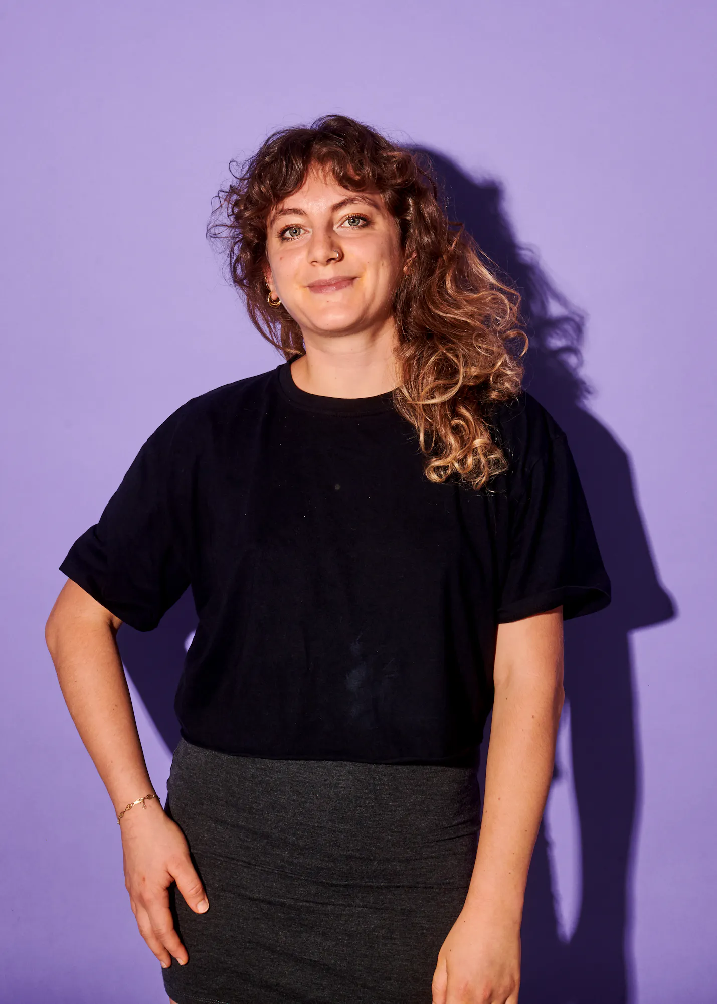 Eine Frau in einem schwarzen T-Shirt und einem grauen Rock steht vor einem lila Hintergrund
