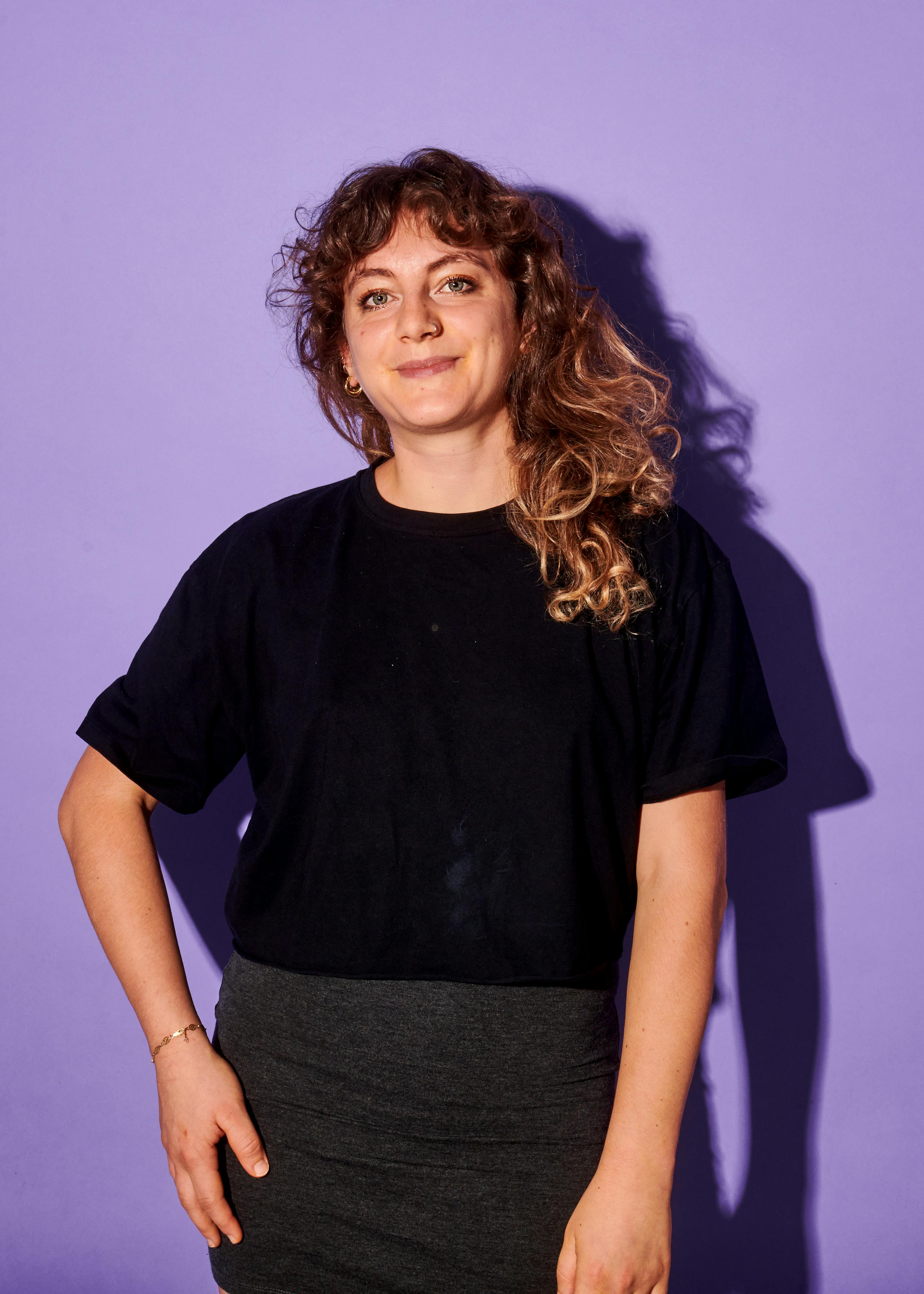 une femme vêtue d' un t-shirt noir et d' une jupe grise se tient devant un fond violet