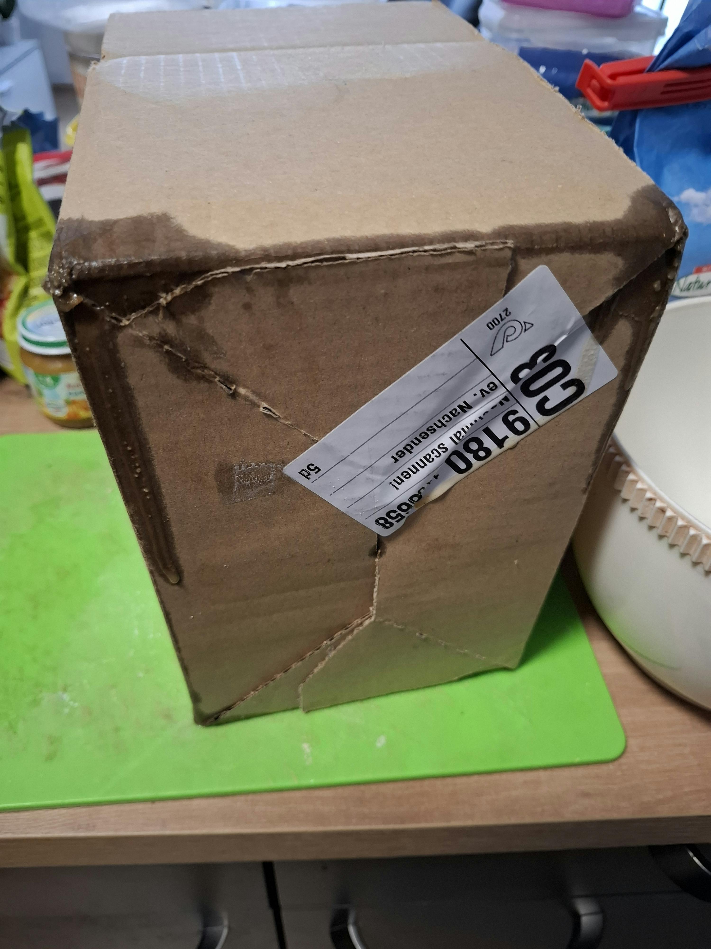 ein nasser, beschädigter karton mit einem versandetikett liegt auf einer holzoberfläche neben einer grünen matte.