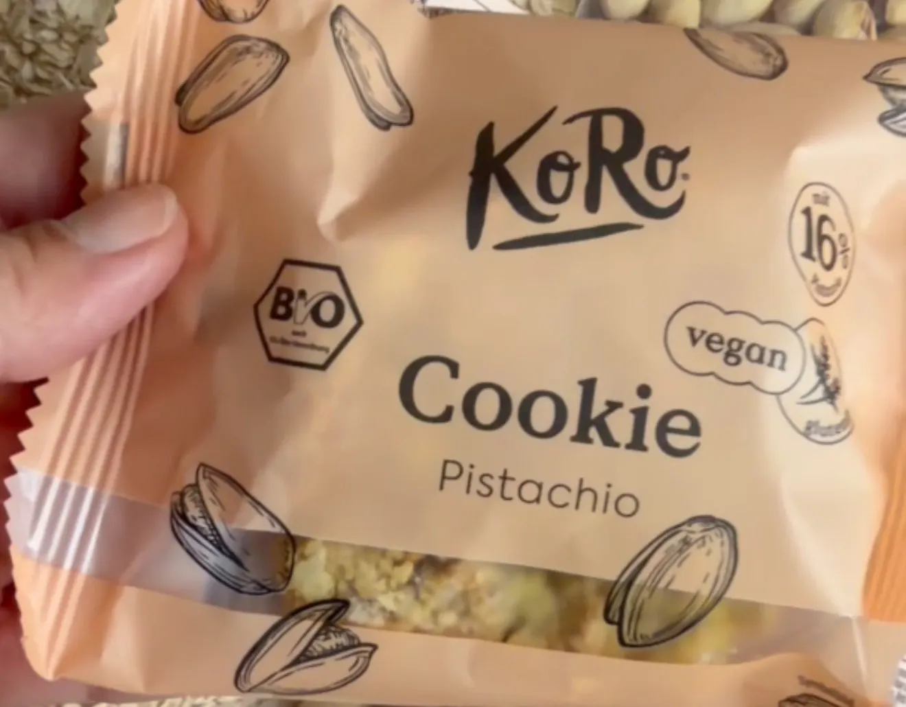eine hand hält eine verpackung eines koro bio-pistazien-cookies, die mit pistazienmotiven verziert ist.