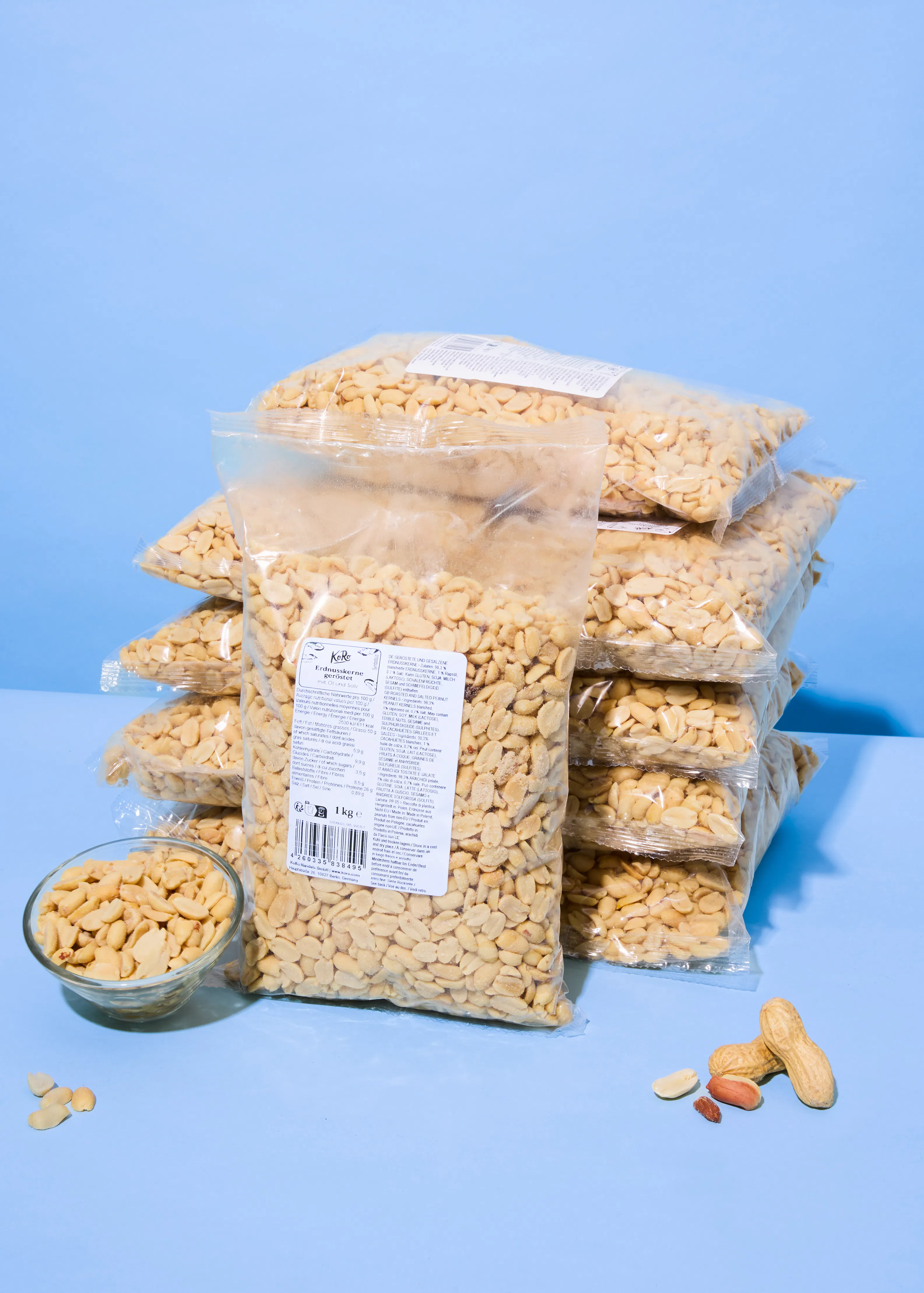 plusieurs sacs transparents de cacahuètes décortiquées sont empilés sur une surface bleue, avec un bol de cacahuètes et quelques cacahuètes éparpillées à côté.