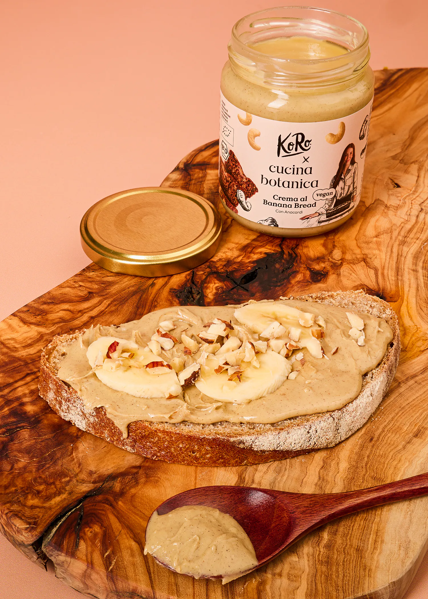 ein glas koro cucina botanica bananenbrot-creme steht neben einer mit der creme bestrichenen brotscheibe, die mit bananenscheiben und gehackten nüssen belegt ist, sowie einem holzlöffel mit etwas creme.