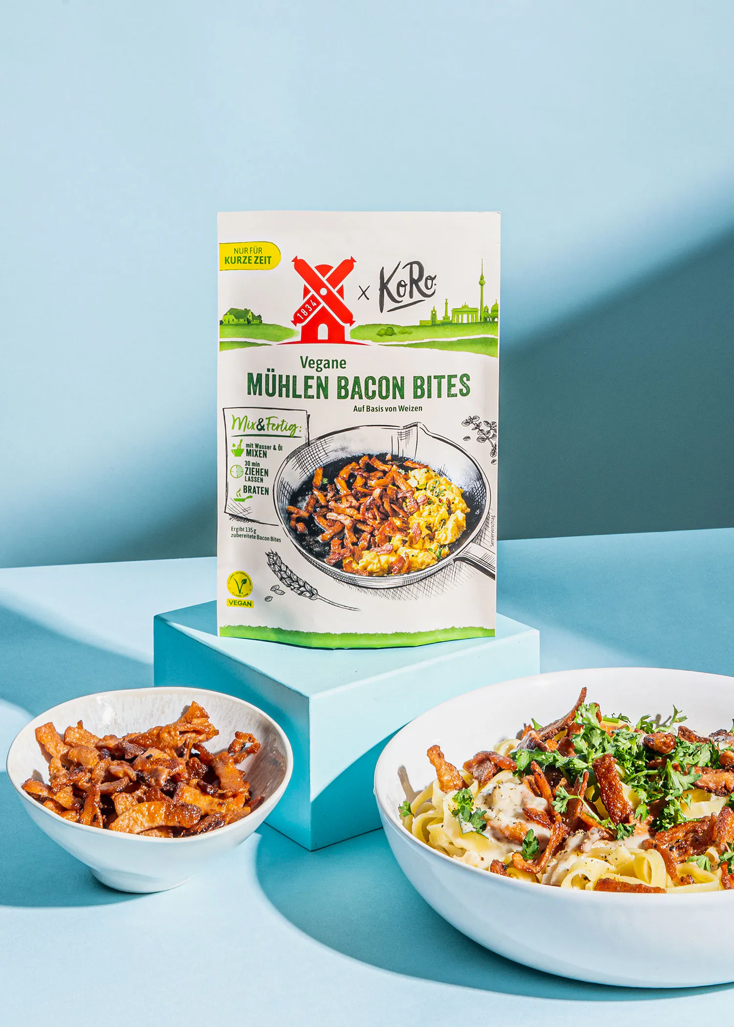 a package of vegane muhlen bacon bites sits next to a bowl of pasta