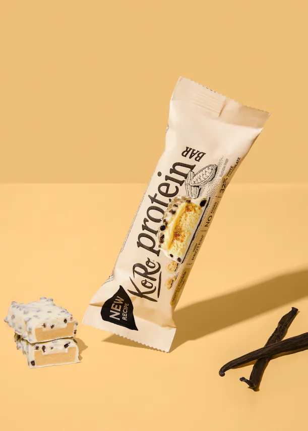 a koro protein bar in vanilla cookie flavor with white chocolate coating is displayed next to two unwrapped bar pieces and two vanilla beans on a yellow background.