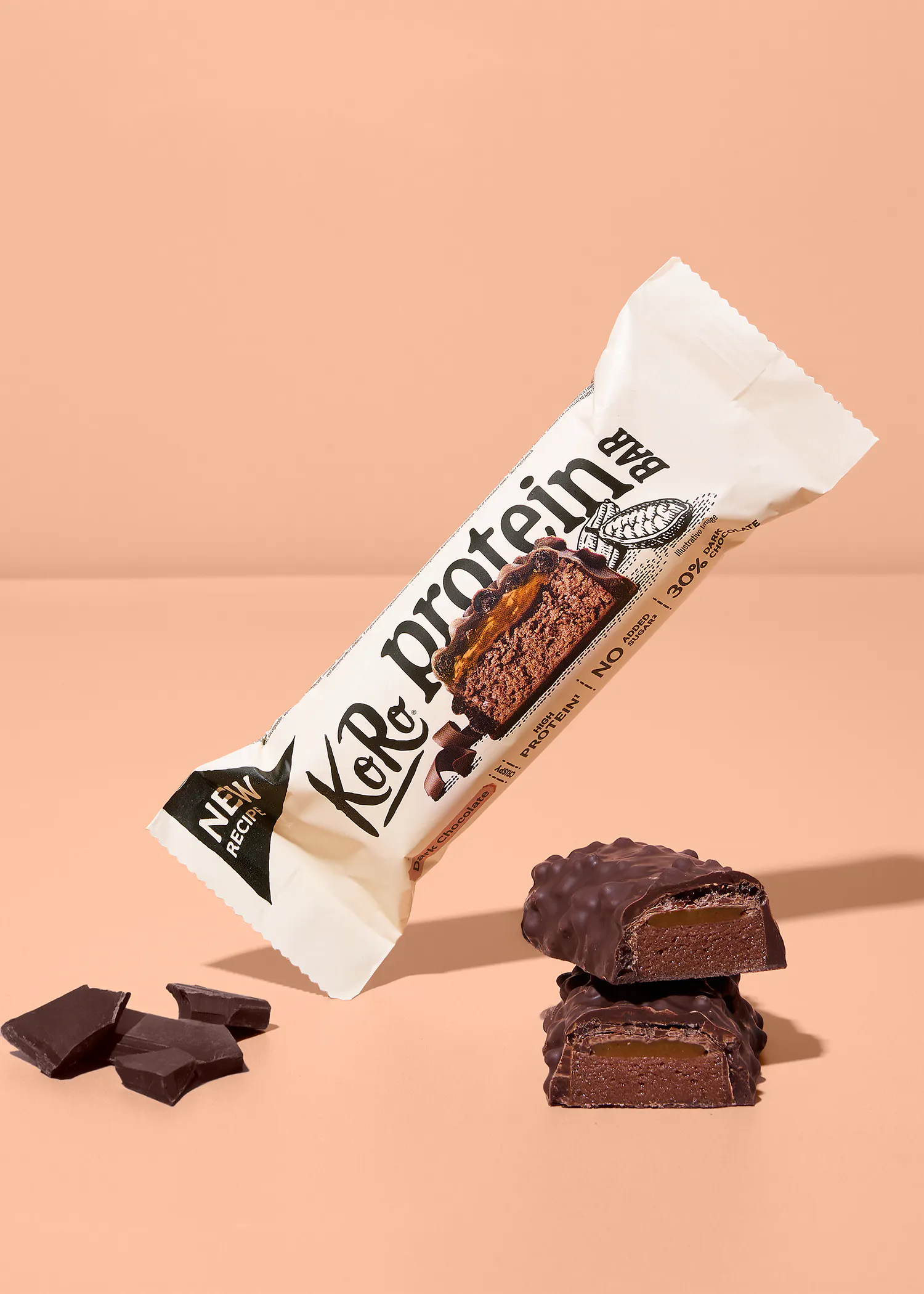 ein koro proteinriegel in der geschmacksrichtung dunkle schokolade mit karamellkern, der neben zwei aufgeschnittenen riegelstücken und schokoladenstücken auf einem beigen hintergrund schwebt.