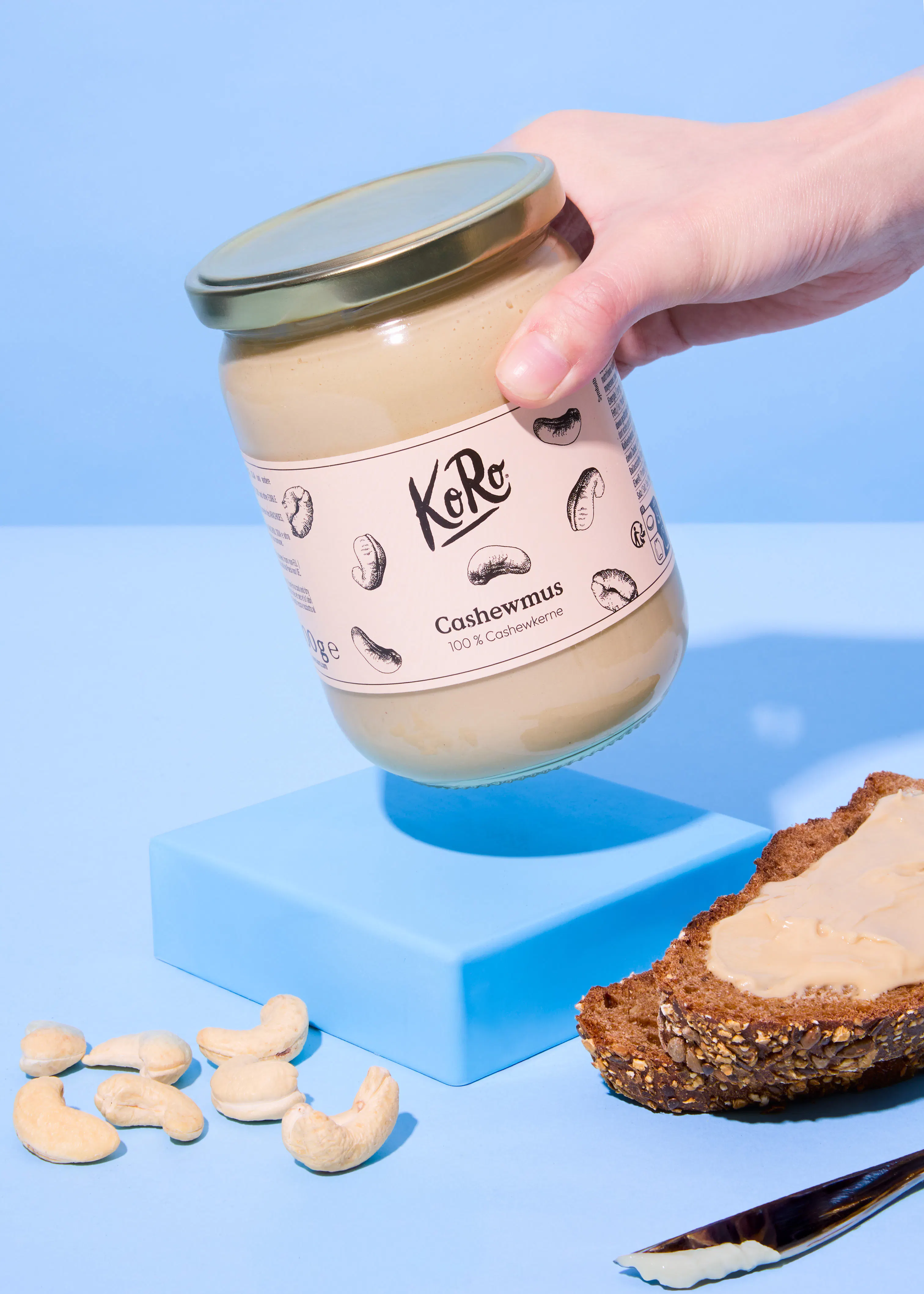 une main tient un pot de purée de cajou koro au-dessus d'une tranche de pain tartinée, de noix de cajou et d'un couteau sur une surface bleue.