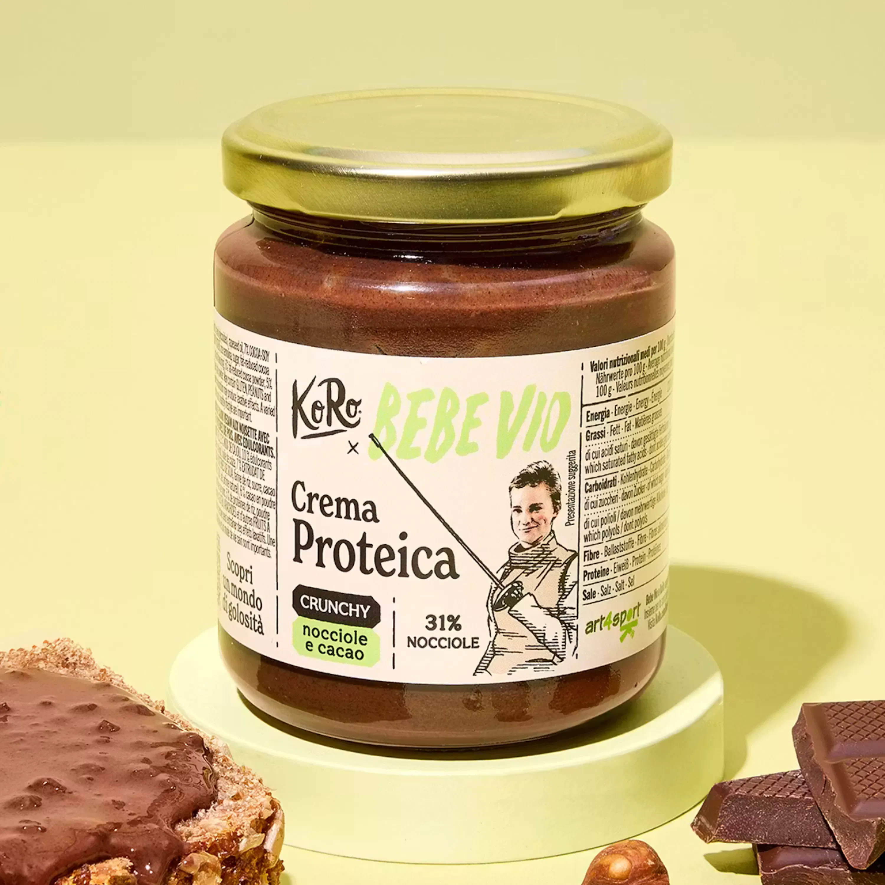 un barattolo di crema proteica croccante alle nocciole e cacao è mostrato su uno sfondo giallo, con una fetta di pane spalmata, pezzi di cioccolato e nocciole sparse.