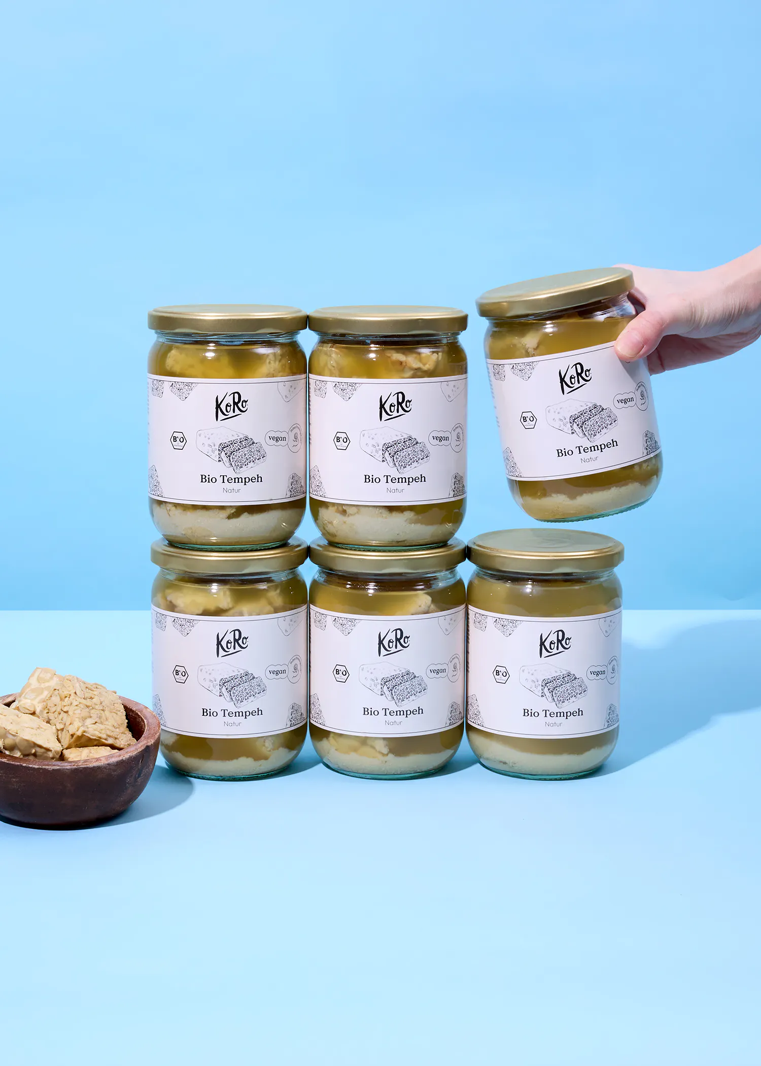 sechs gläser koro bio tempeh natur, eines davon wird von einer hand gehalten, und eine schale mit tempeh-stücken auf blauem hintergrund.