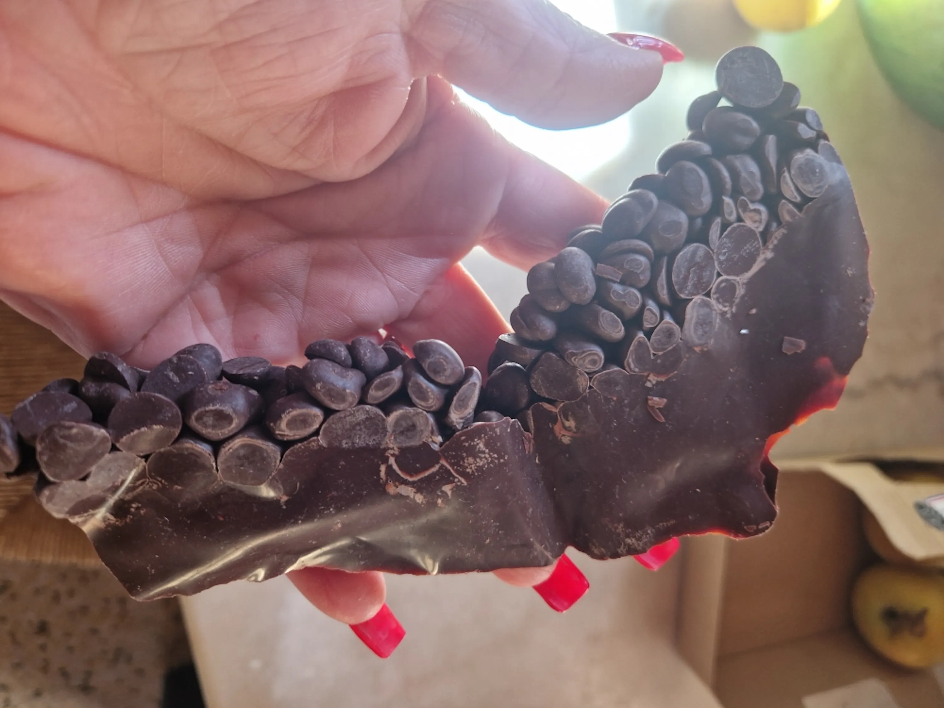 a hand with red nail polish holds a piece of dark chocolate bark covered in chocolate chips.