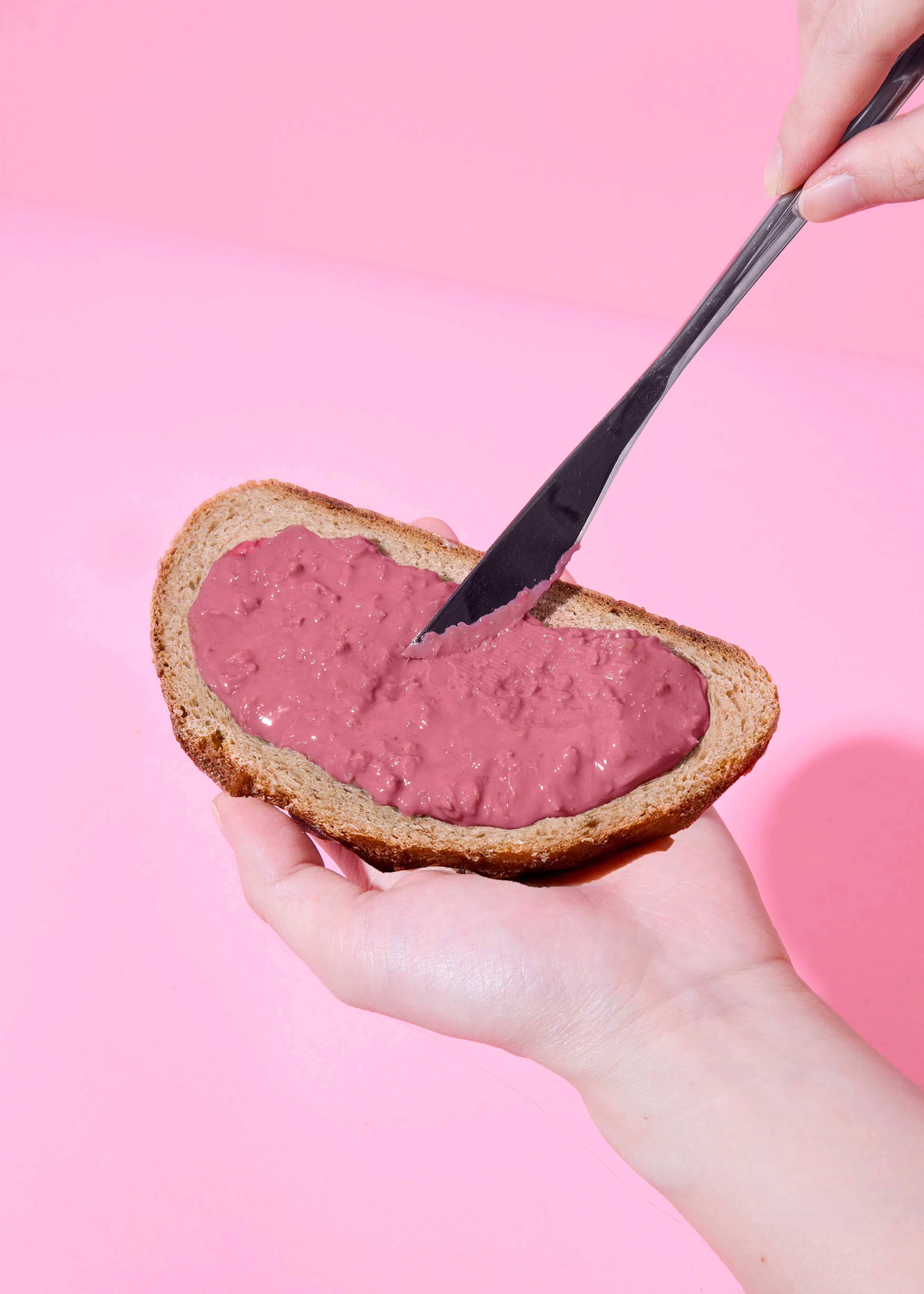 eine hand hält eine scheibe brot mit rosa aufstrich, der mit einem messer auf einem rosa hintergrund verteilt wird.