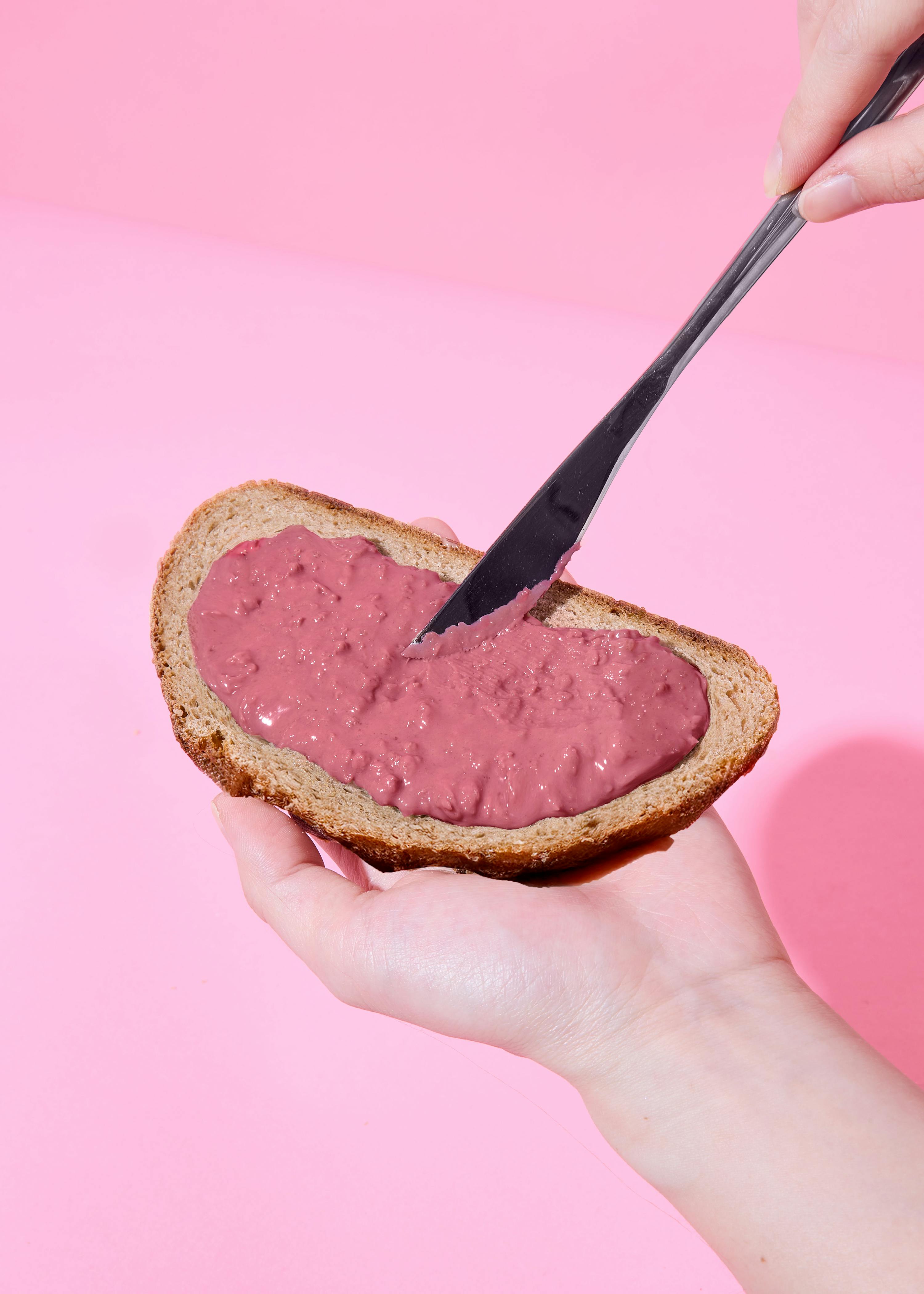 eine hand hält eine scheibe brot mit rosa aufstrich, der mit einem messer auf einem rosa hintergrund verteilt wird.