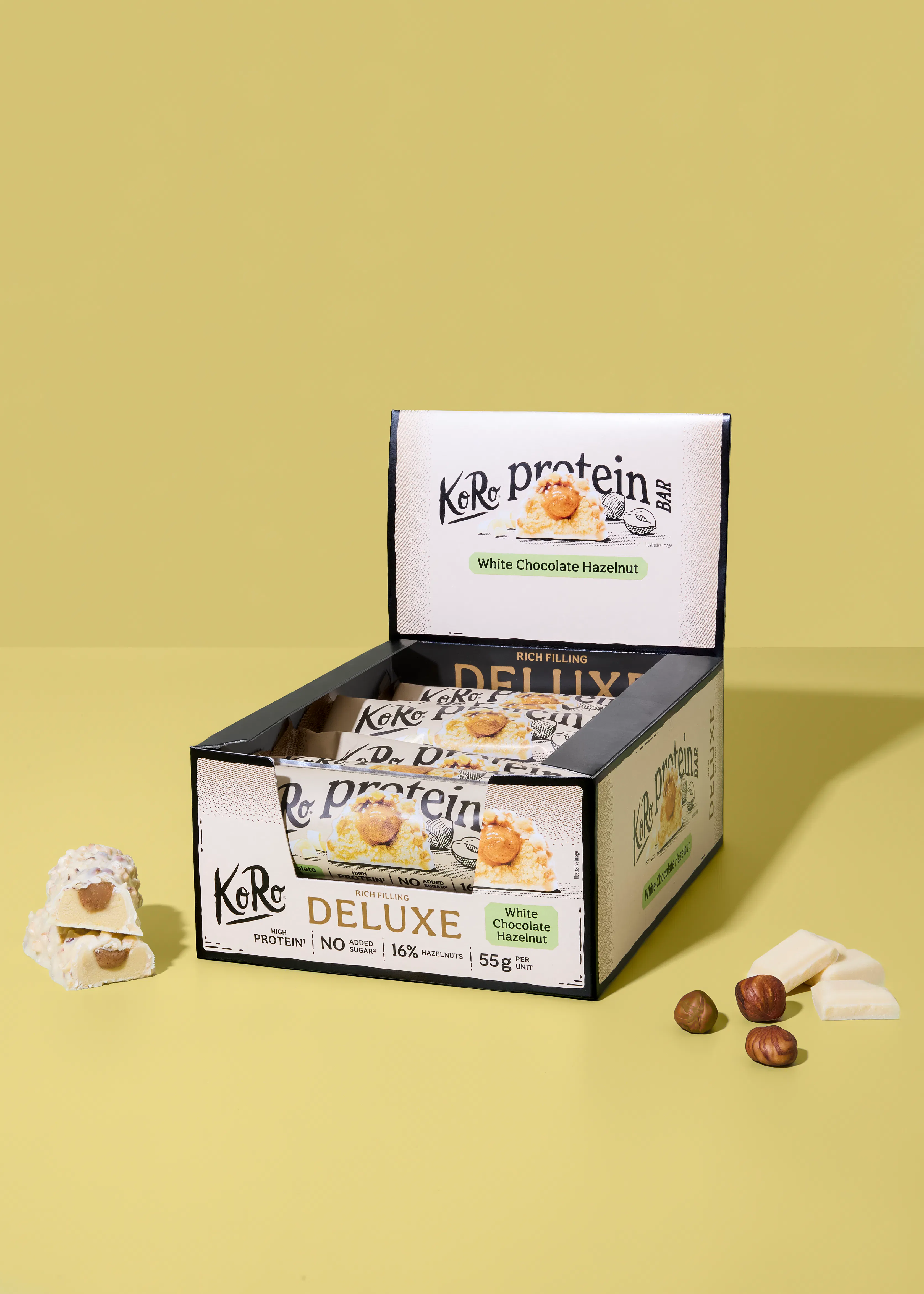 une boîte ouverte de barres protéinées koro deluxe chocolat blanc noisette, avec une barre coupée montrant le fourrage et des noisettes et morceaux de chocolat blanc à côté sur un fond jaune.
