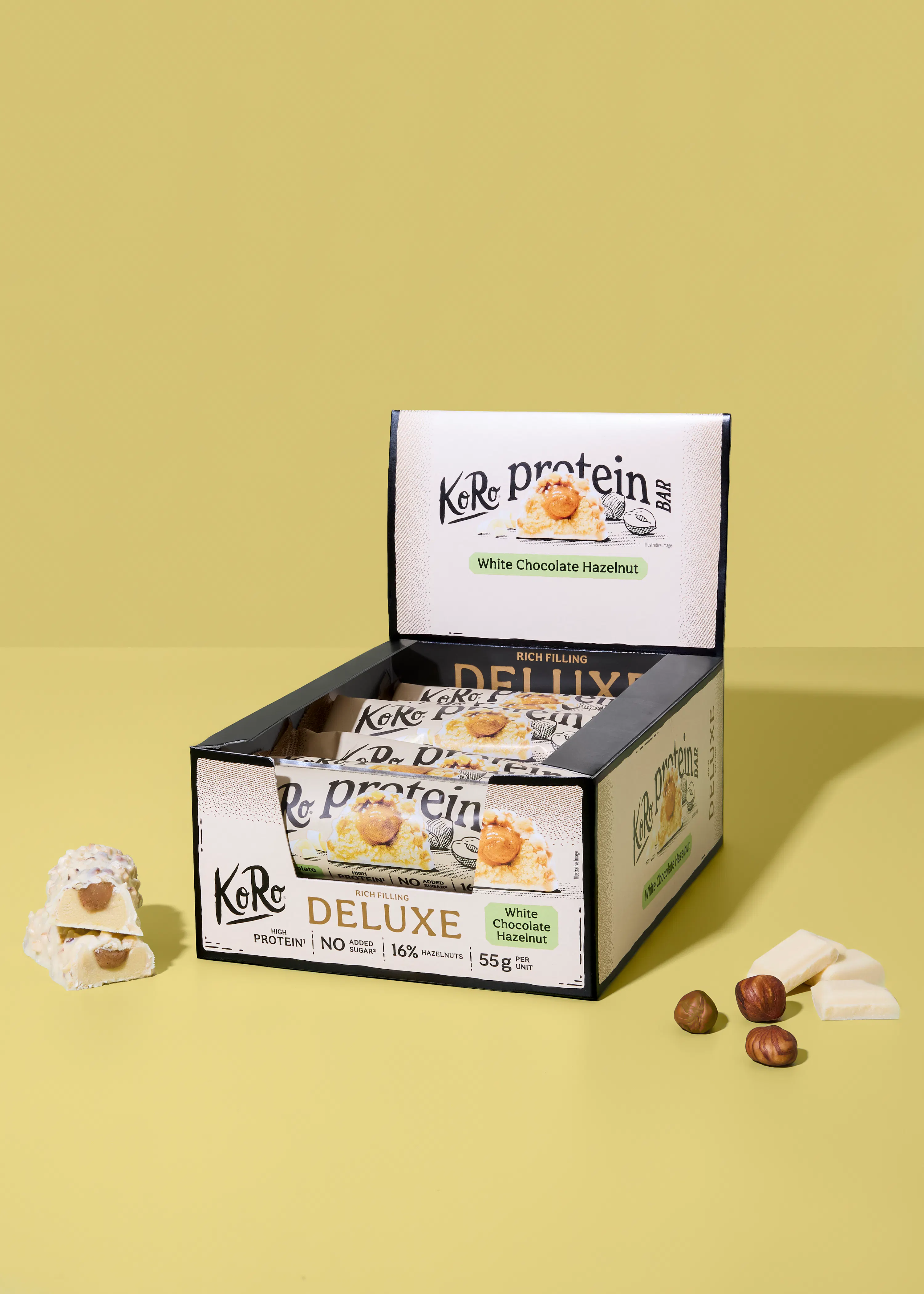 ein offener displaykarton mit koro protein deluxe white chocolate hazelnut riegeln, daneben ein angeschnittener riegel, lose haselnüsse und weiße schokoladenstücke auf gelbem hintergrund.