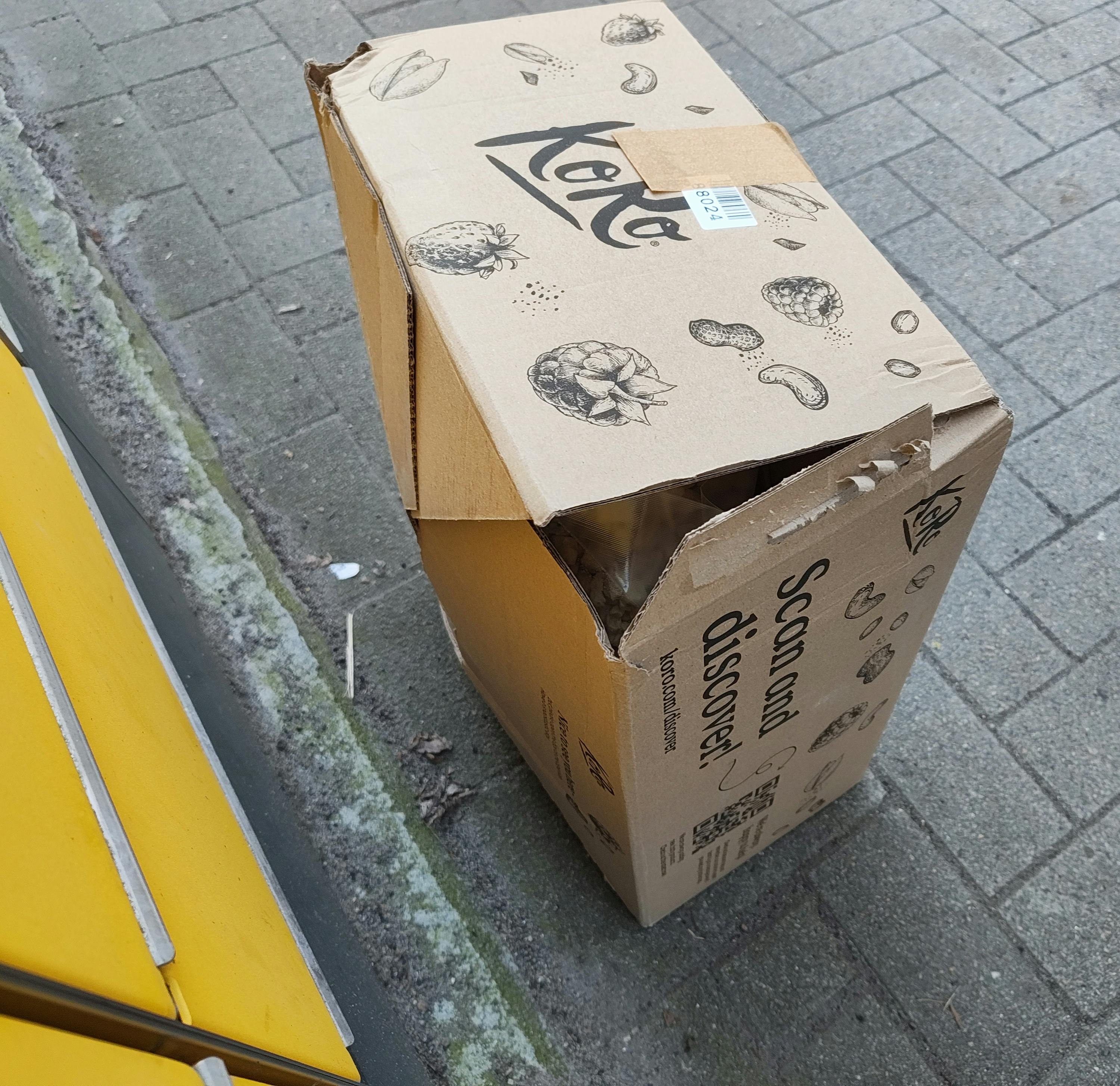 ein teilweise geöffneter koro-karton mit abbildungen von nüssen und früchten steht auf einem gepflasterten weg neben einem gelben objekt.