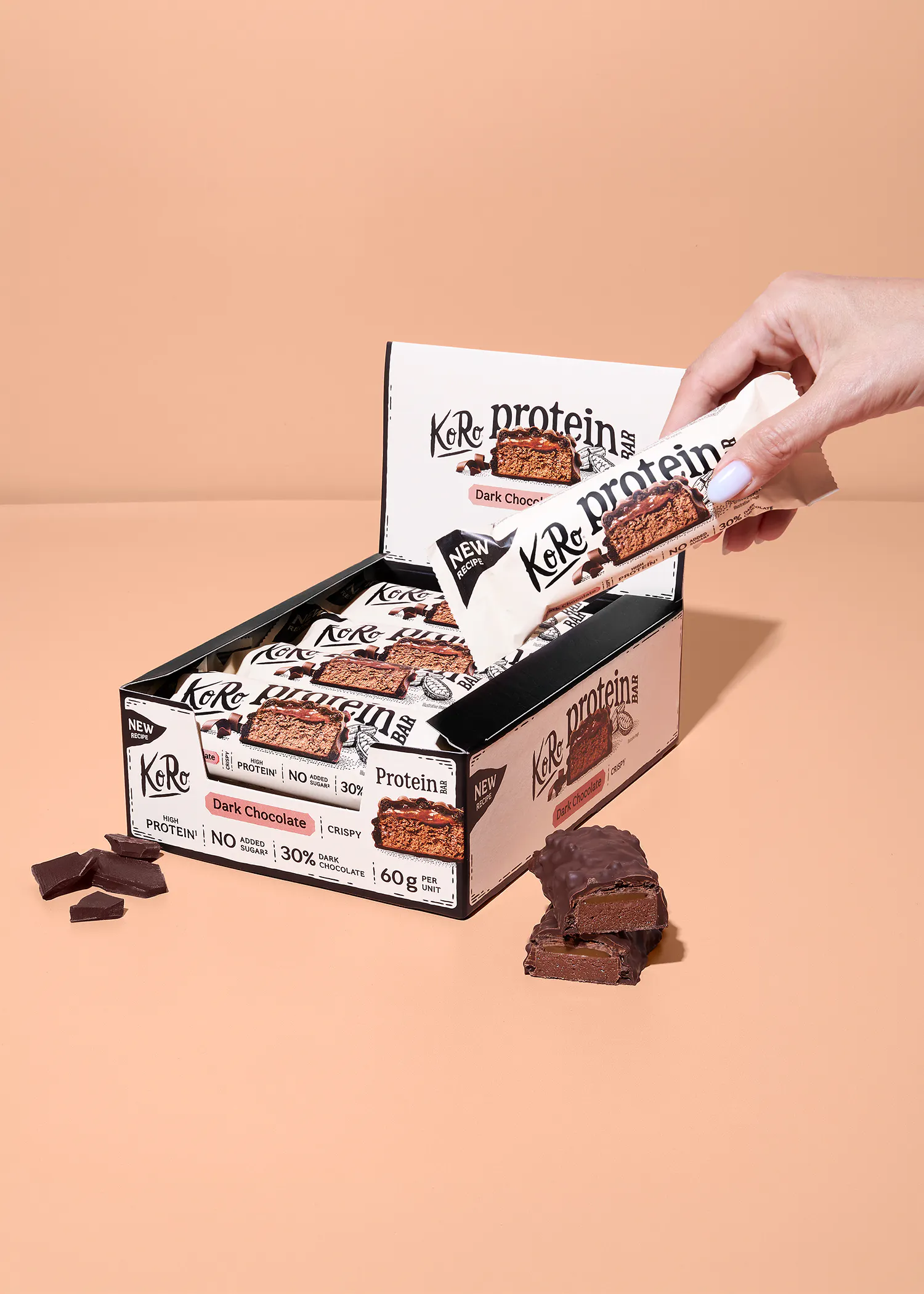 eine hand hält einen koro proteinriegel mit dunkler schokolade über einer offenen verkaufsbox voller weiterer riegel vor einem beigen hintergrund.