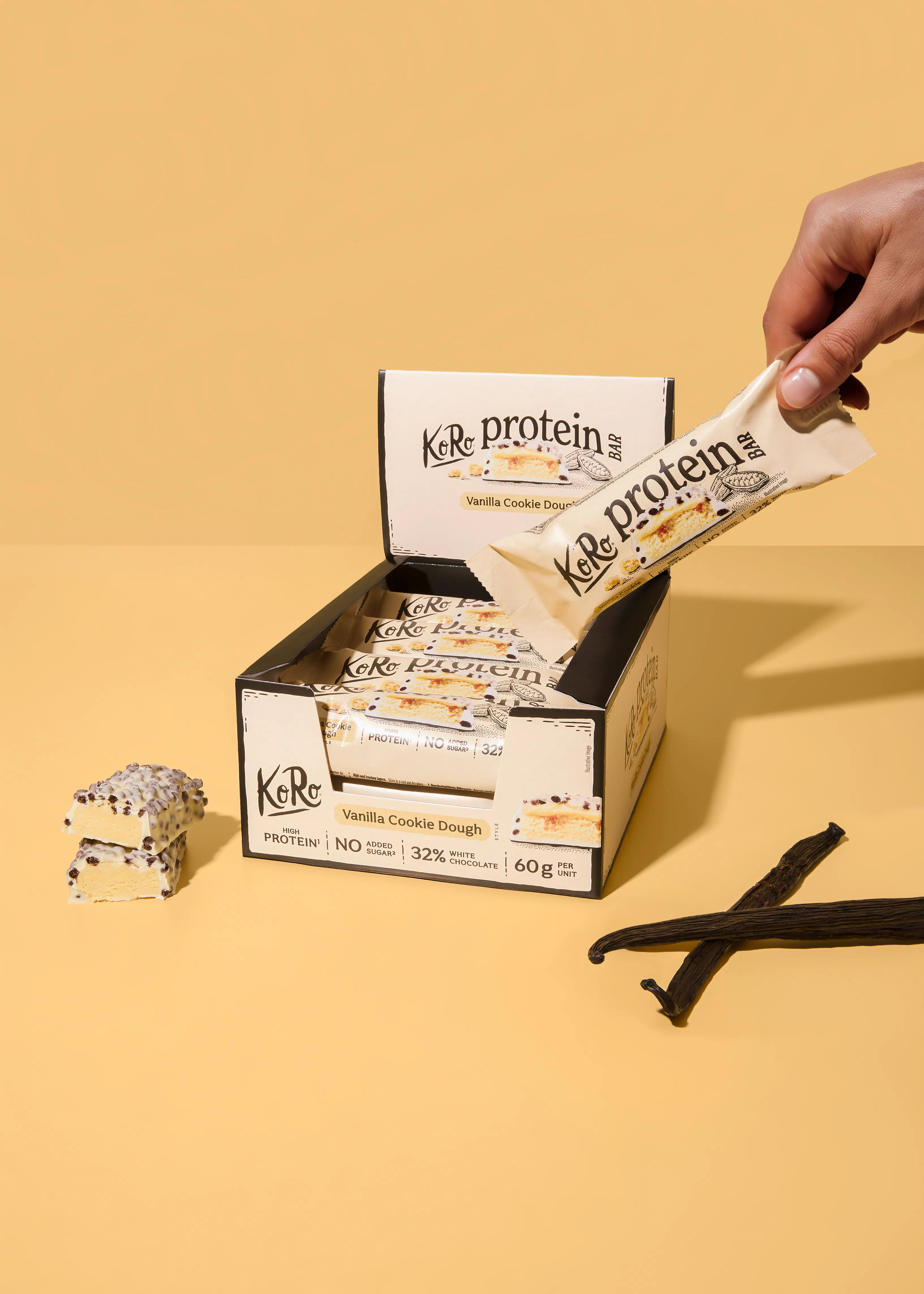 eine hand nimmt einen vanille-cookie-dough-proteinriegel aus einer offenen schachtel, neben der zwei halbierte riegel und zwei vanilleschoten auf gelbem hintergrund liegen.